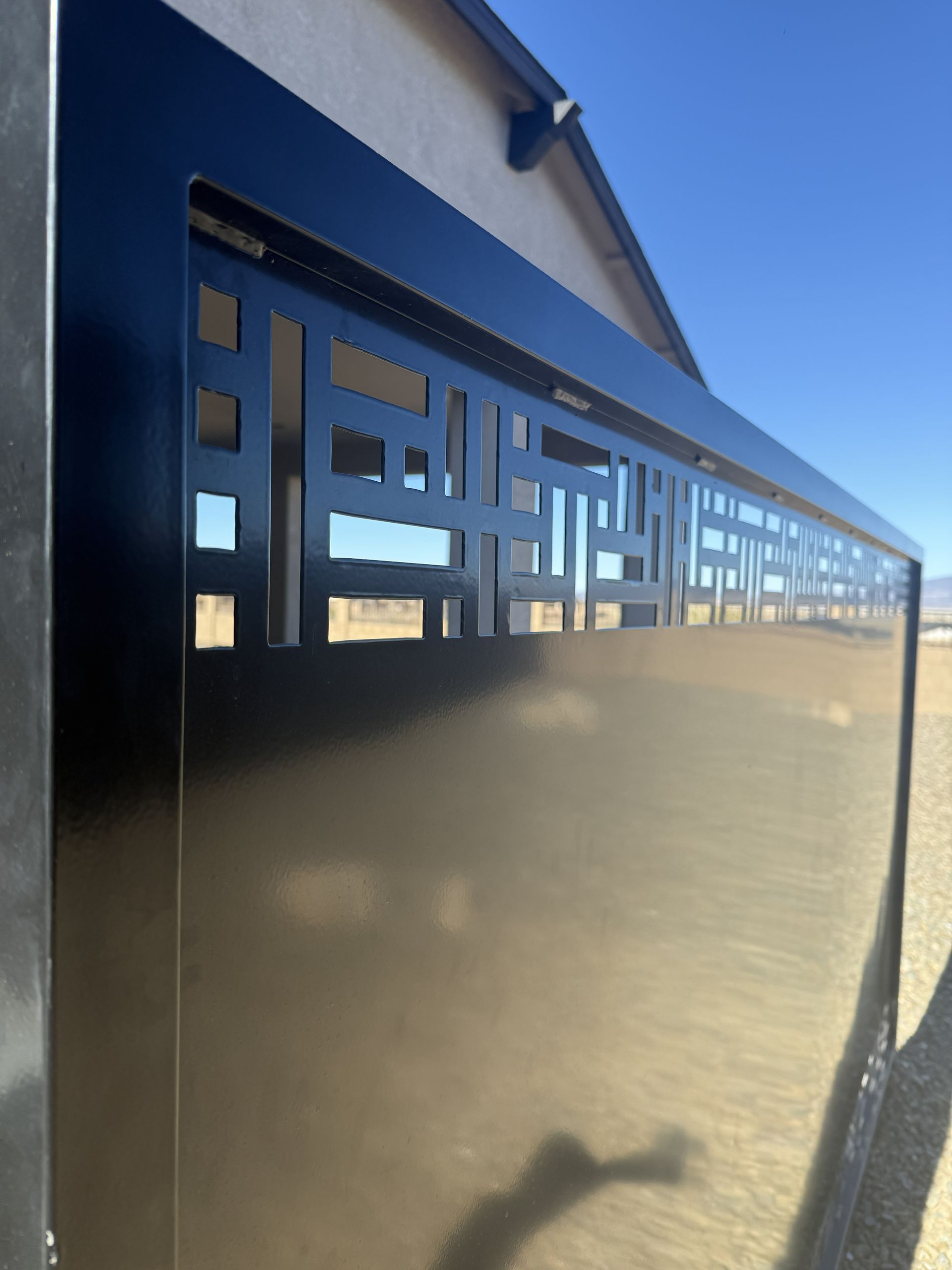 A black metal gate with a geometric, laser-cut pattern at the top, set against a desert landscape and blue sky.