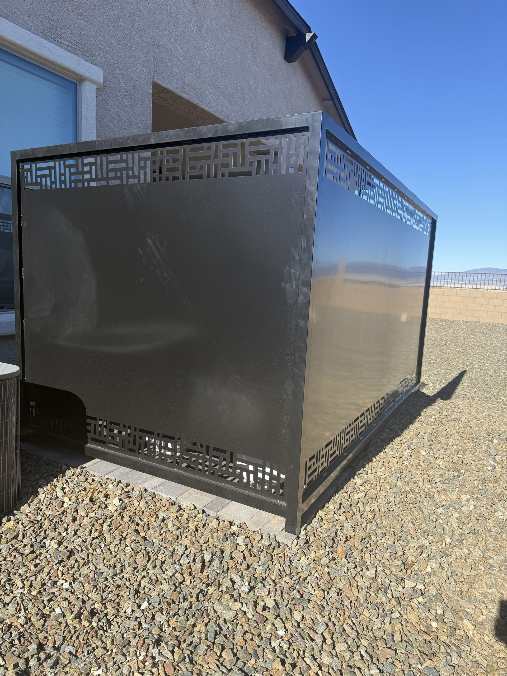 A dark metal privacy screen with geometric laser-cut patterns, installed on gravel next to a building.