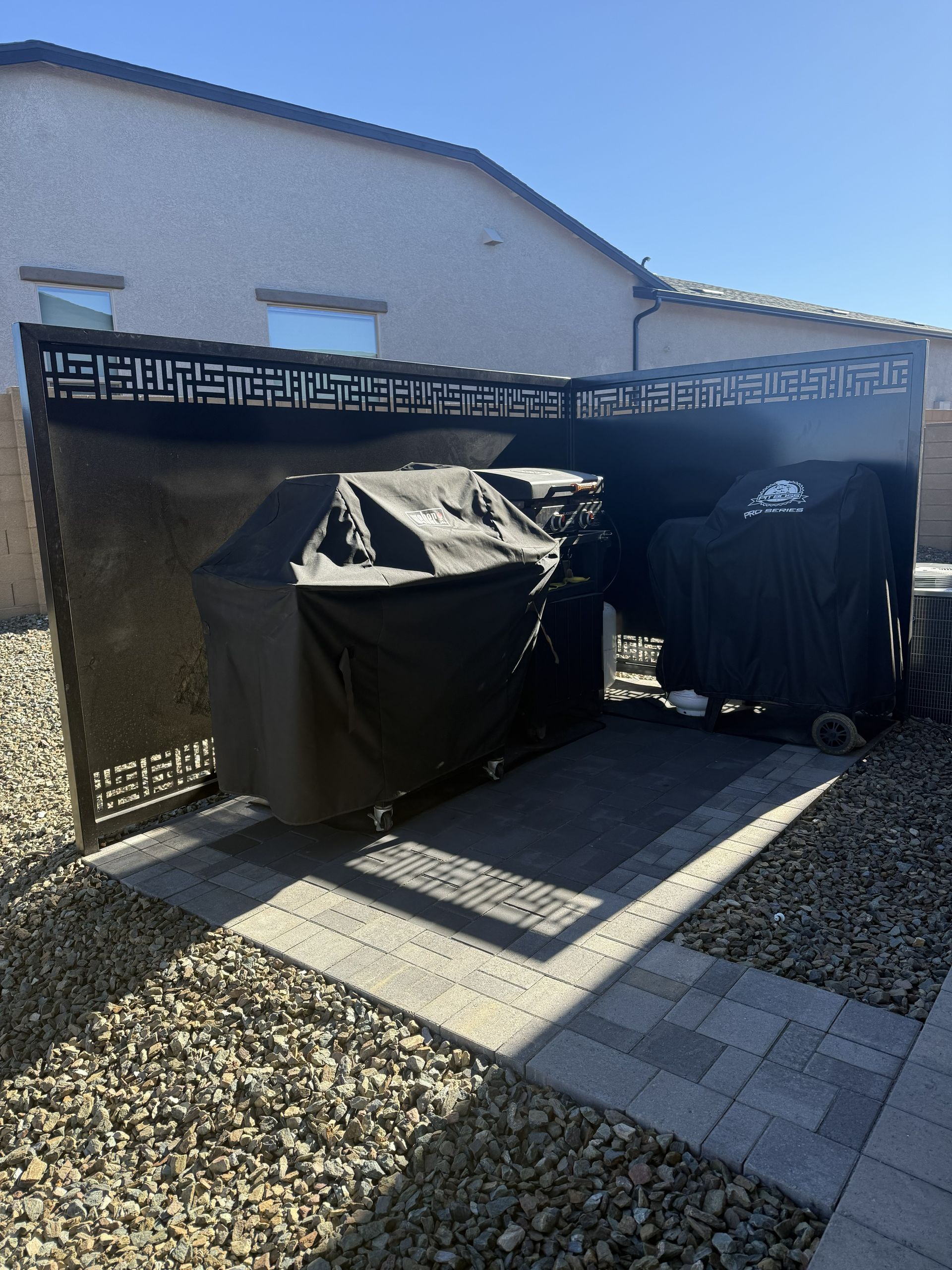 Two grills covered in black tarps sit on a paved patio behind a decorative, dark metal privacy screen outdoors.