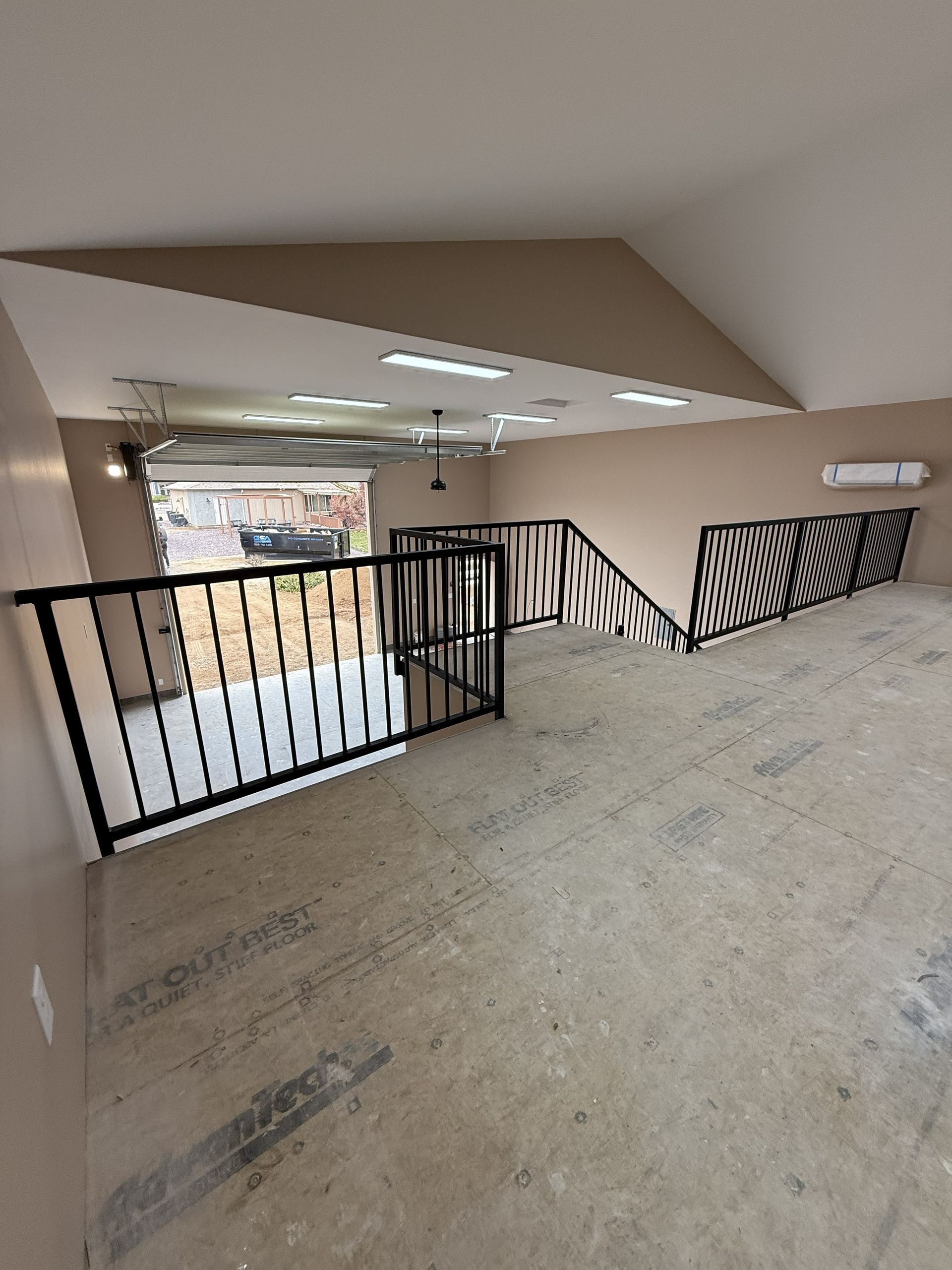 An unfinished loft area with wood subflooring, black metal railings, beige walls, and a view into a garage below.