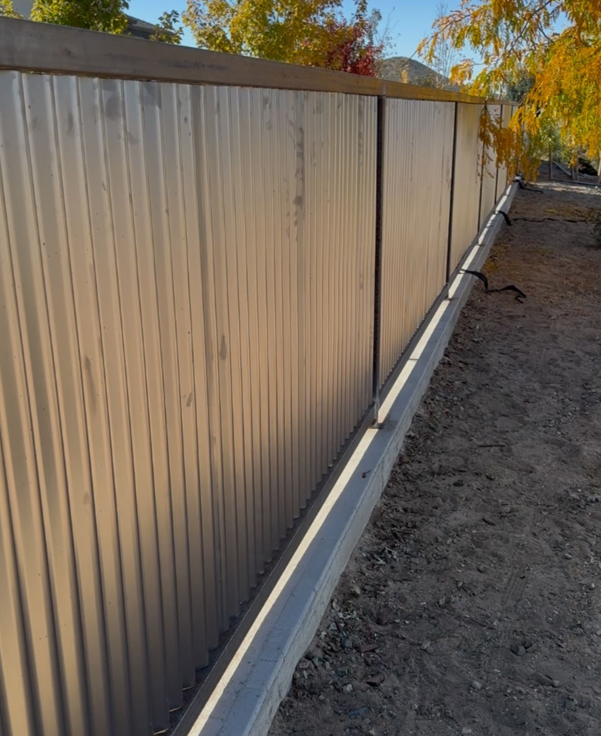 Tan corrugated metal fence with a wooden top, against a dirt ground, illuminated by sunlight.