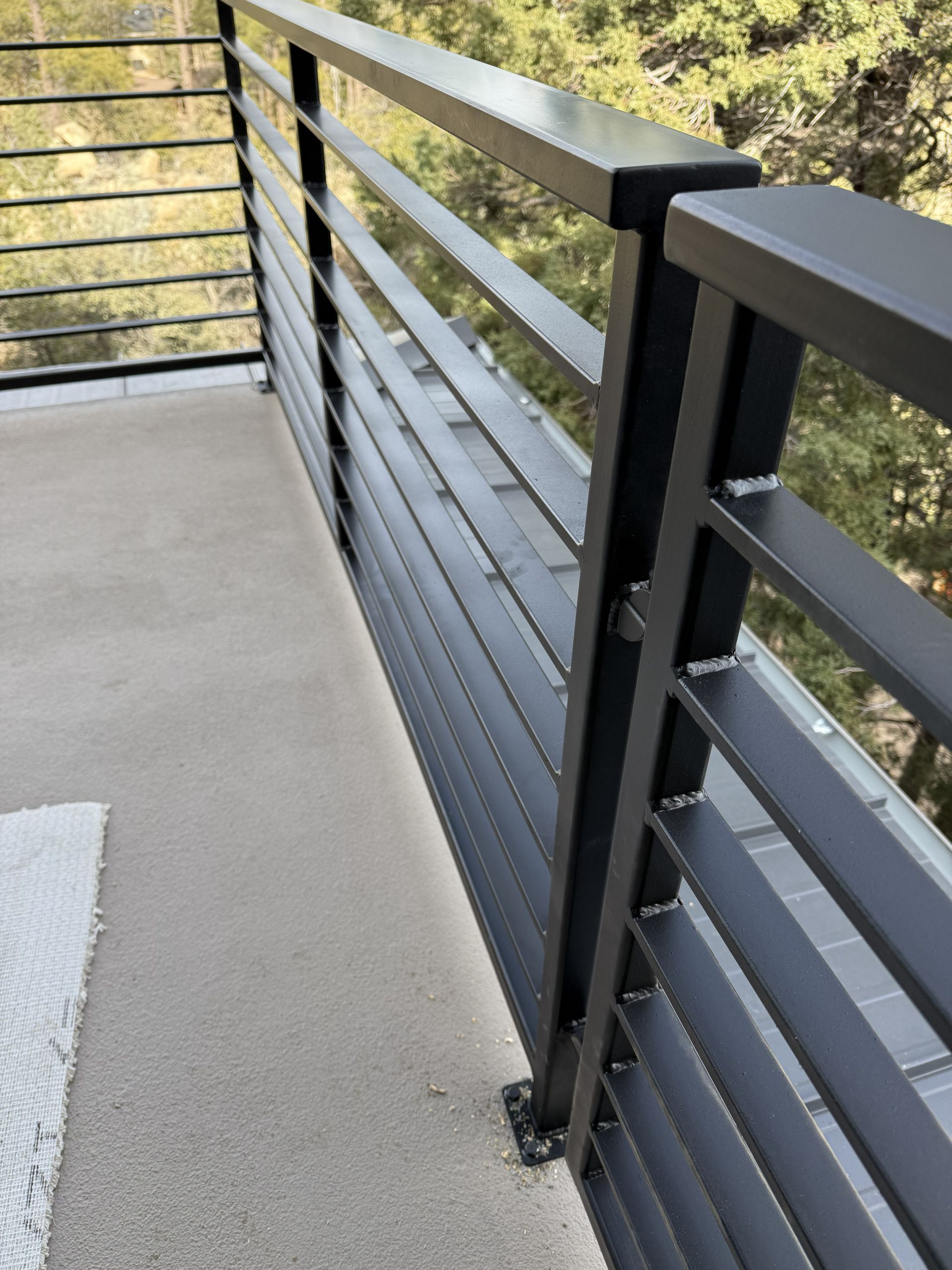 Close-up of a modern matte black metal balcony railing with horizontal slats, overlooking a treed landscape.