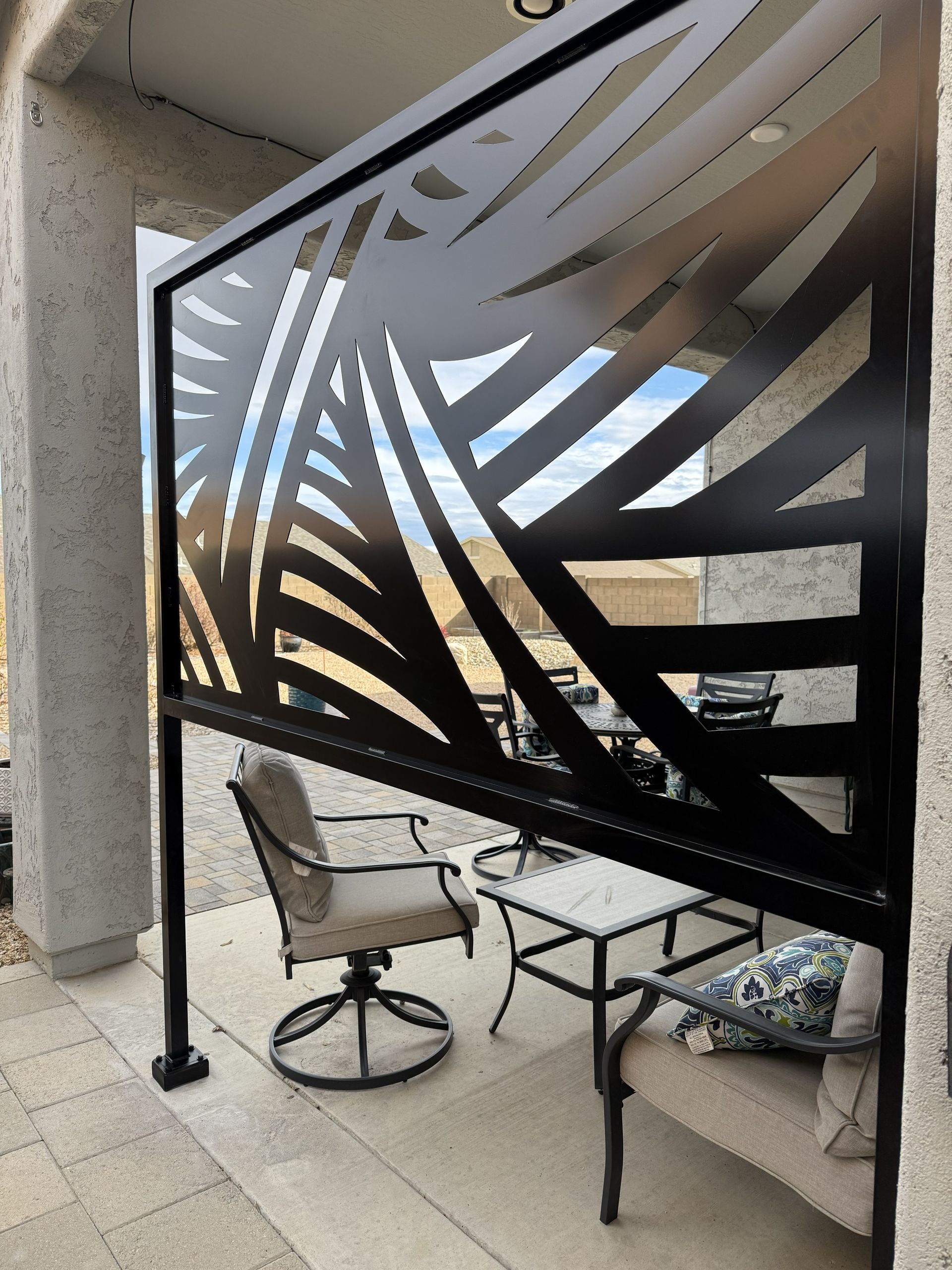 A black metal privacy screen with a palm leaf cutout design stands on a patio next to outdoor furniture.