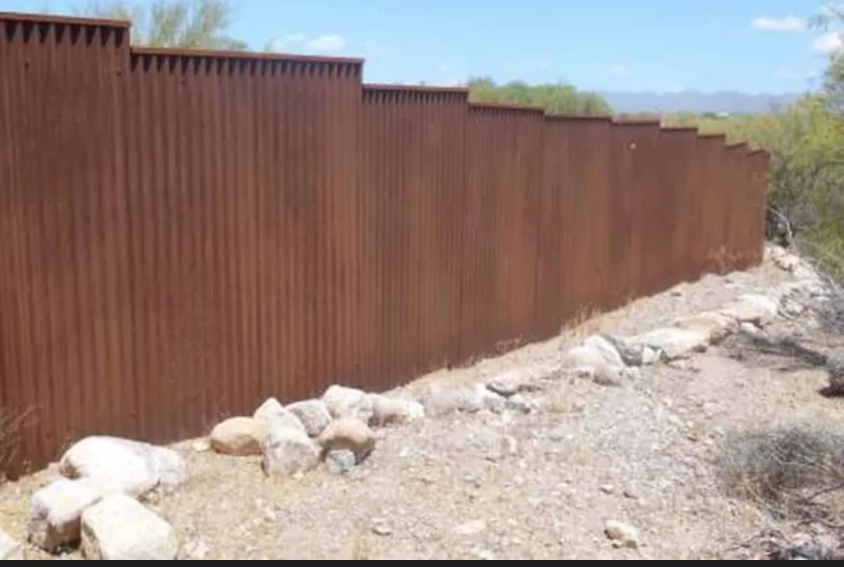 A long, rusted metal border wall spans a rocky, desert landscape under a bright sky.