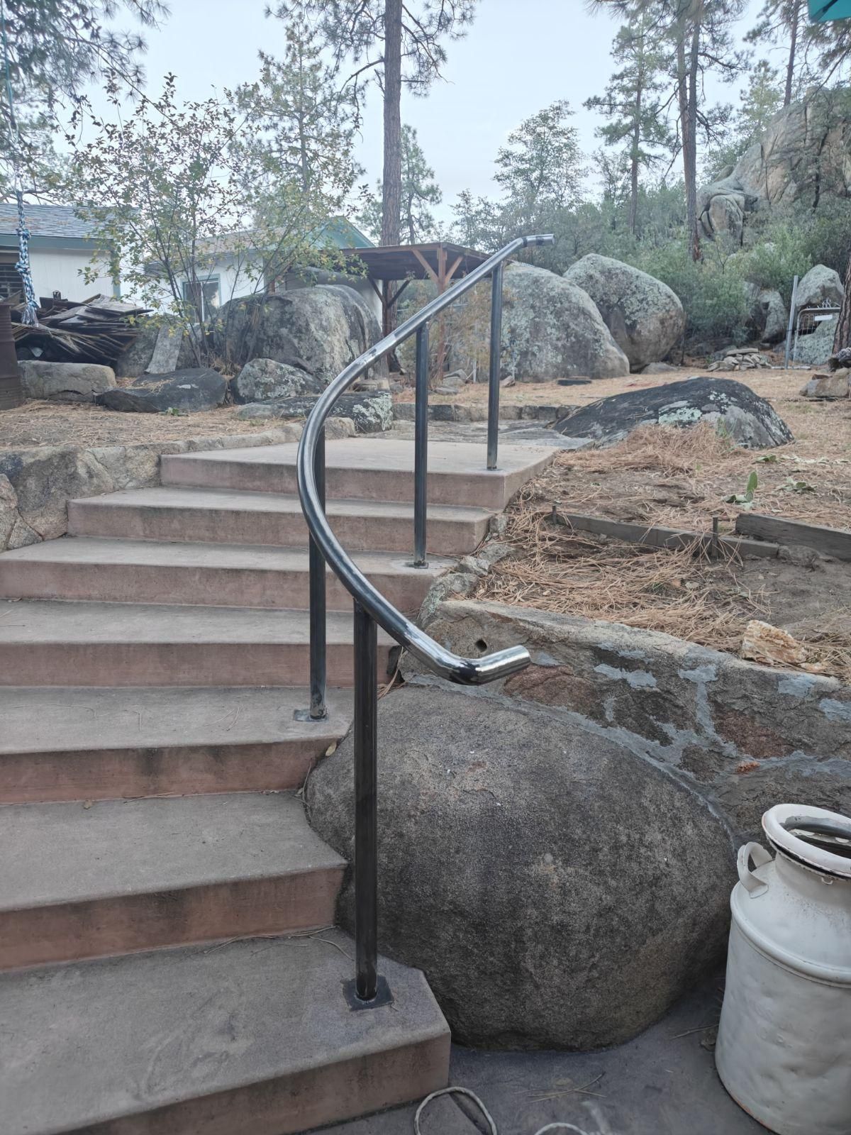 Outdoor concrete stairs with curved metal handrail, leading up to a rocky garden area.