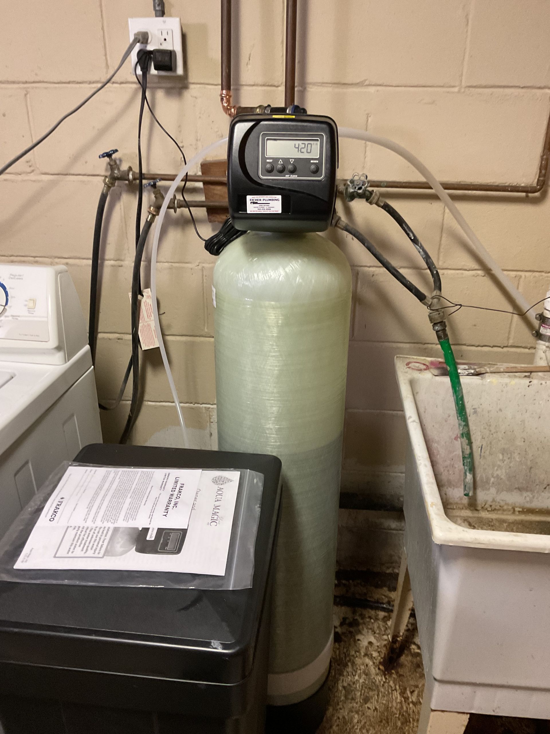 Water softener tank with control head in a laundry/utility room, beside a washing machine and utility sink.