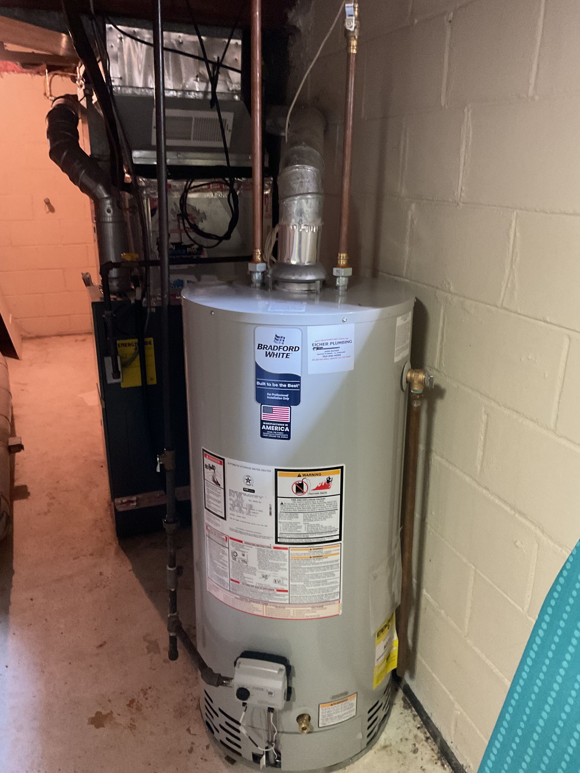 Gray water heater in a narrow basement utility area beside a brick wall and pipes.