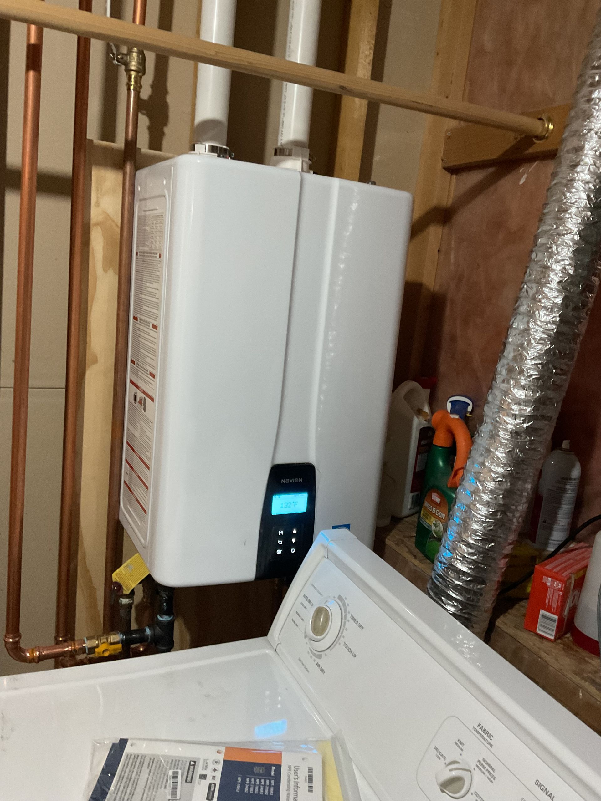 White wall-mounted unit in a utility room beside exposed copper pipes and a washer lid.