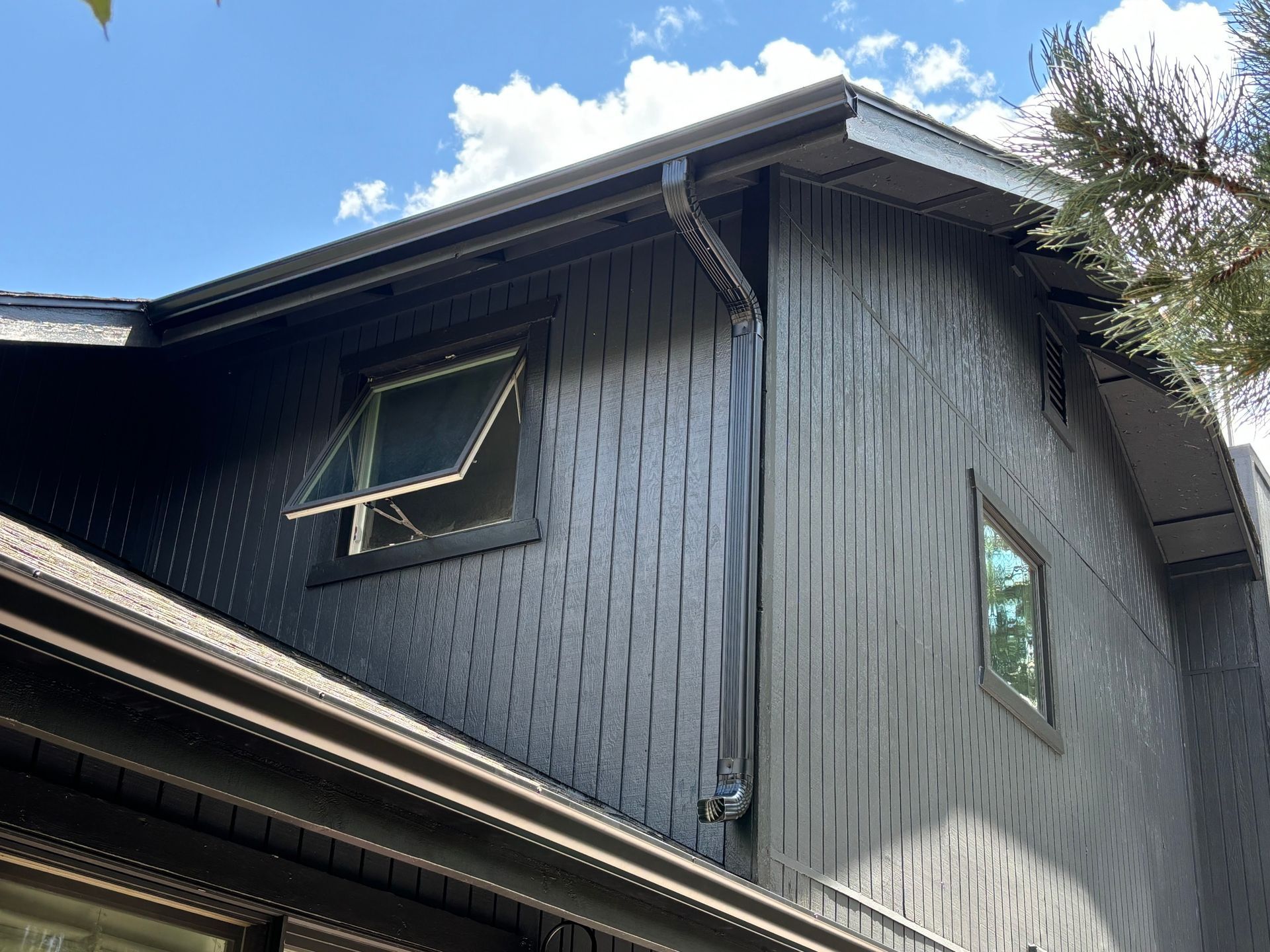 A dark-colored house exterior with textured, vertical siding, a downspout, and two windows against a bright blue sky.