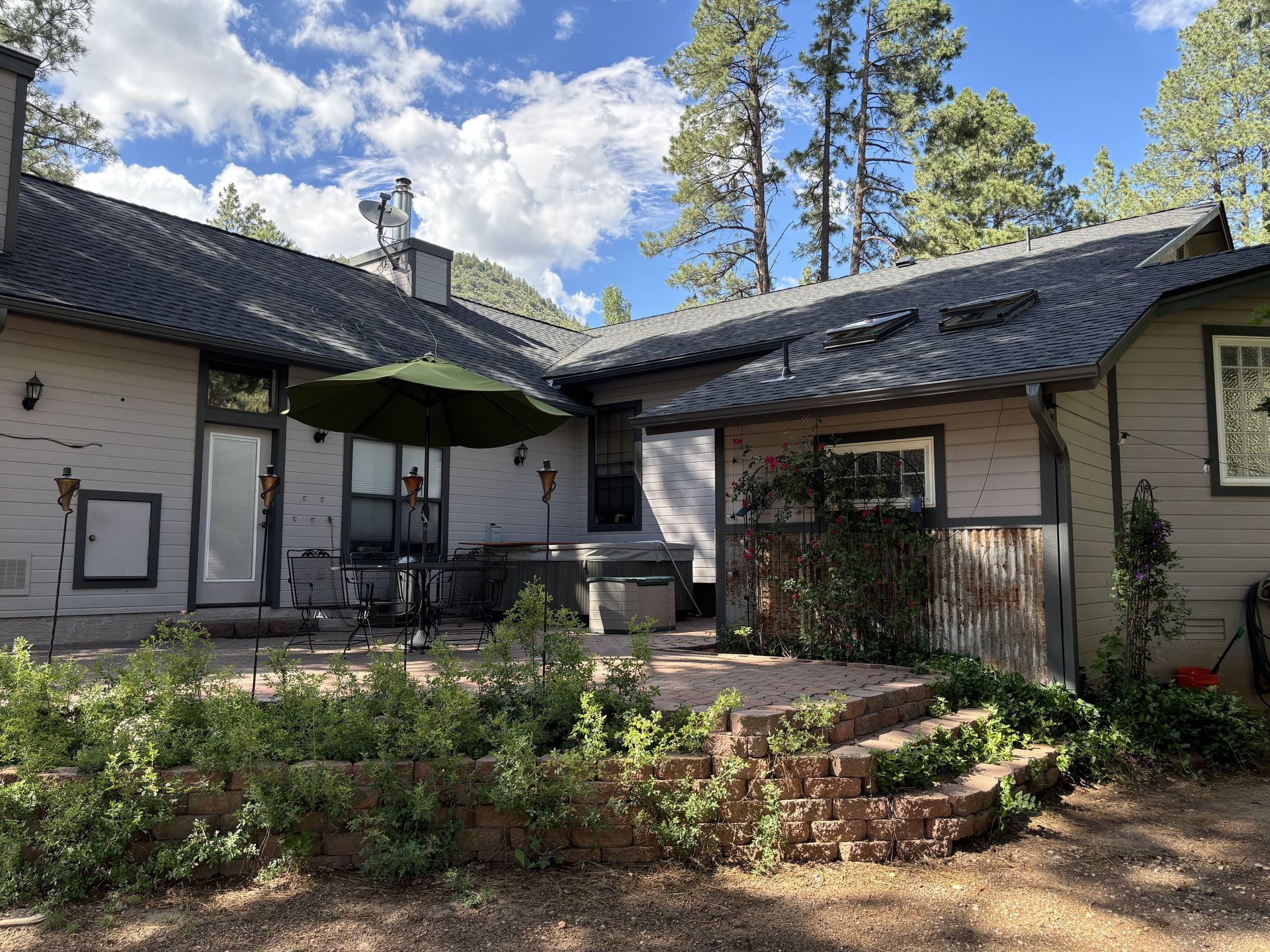 A back patio with a green umbrella, a hot tub, and lush landscaping against a single-story house in a wooded setting.