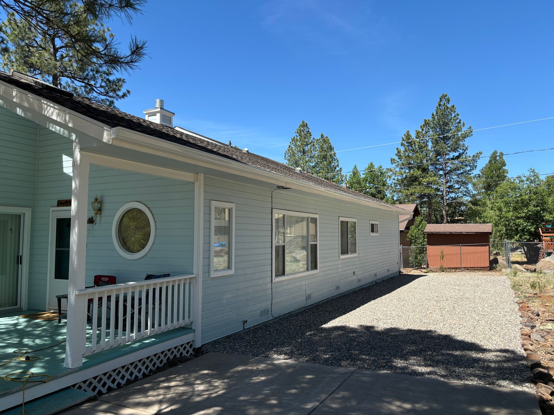 A light blue house with a white front porch railing and trim, featuring a round window, set on a gravel lot under trees.