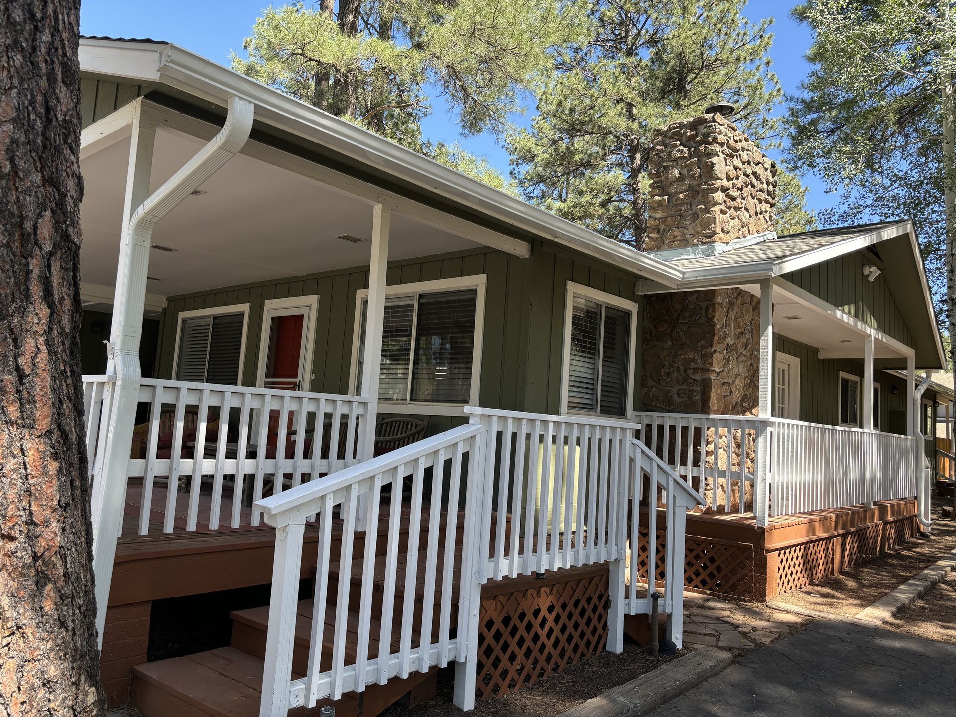 Green cabin exterior with a large front porch, white railings, and a stone chimney nestled among pine trees.