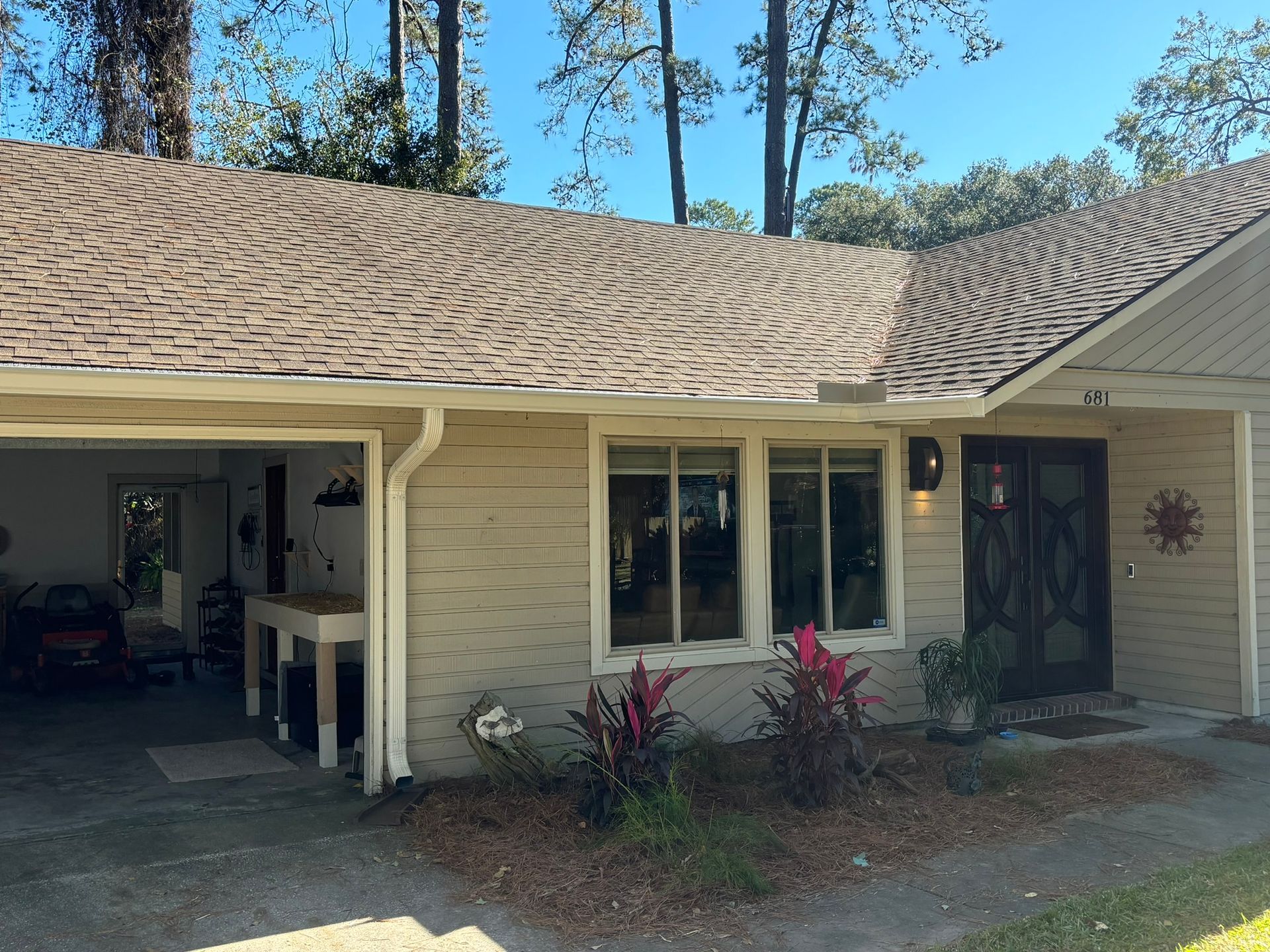 A tan house with a brown shingled roof, double front doors, a wide window, and an open garage, viewed from the driveway.