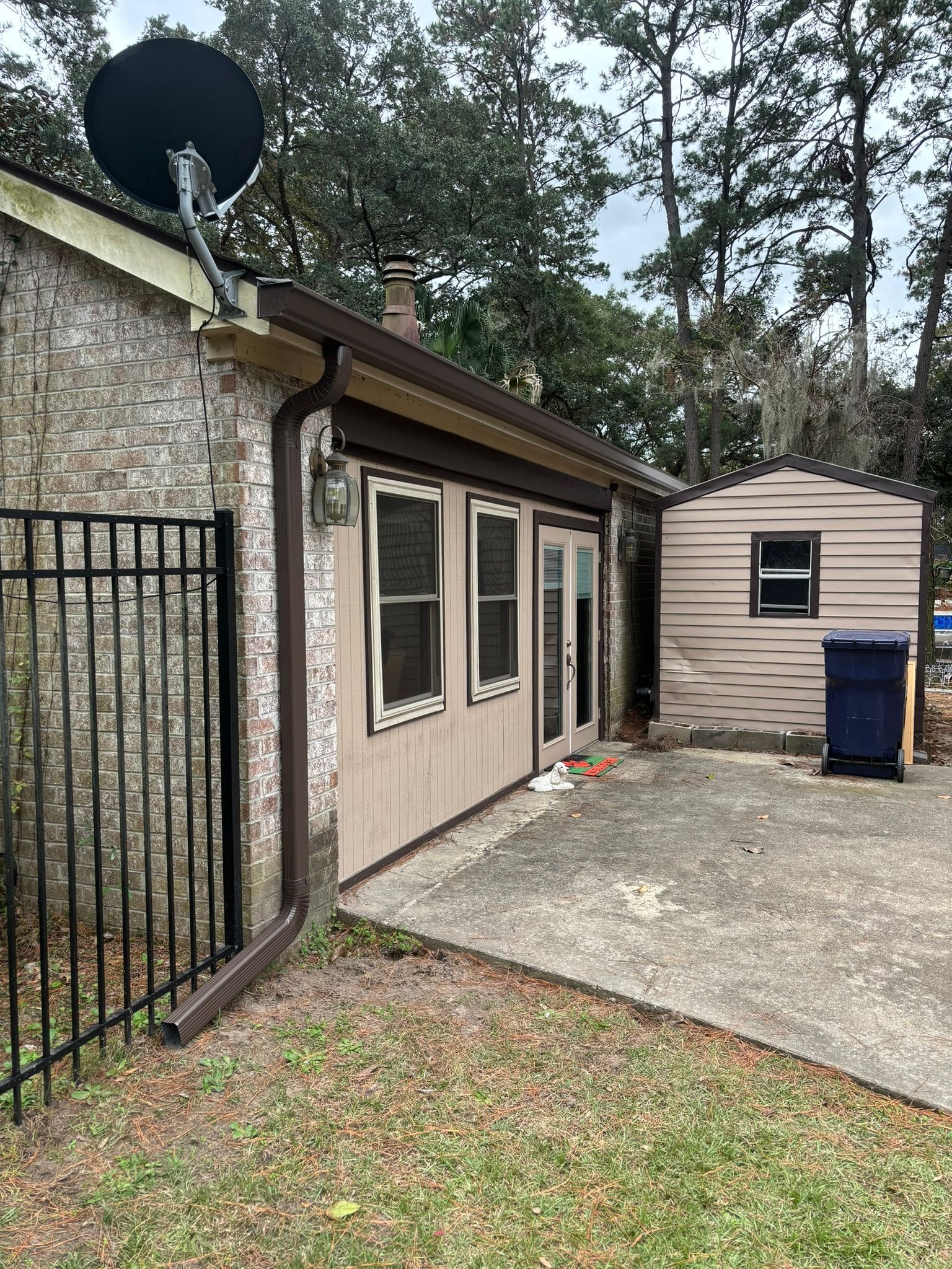 A side view of a brick house with a brown downspout, a satellite dish, a patio door, and a small tan shed on the side.