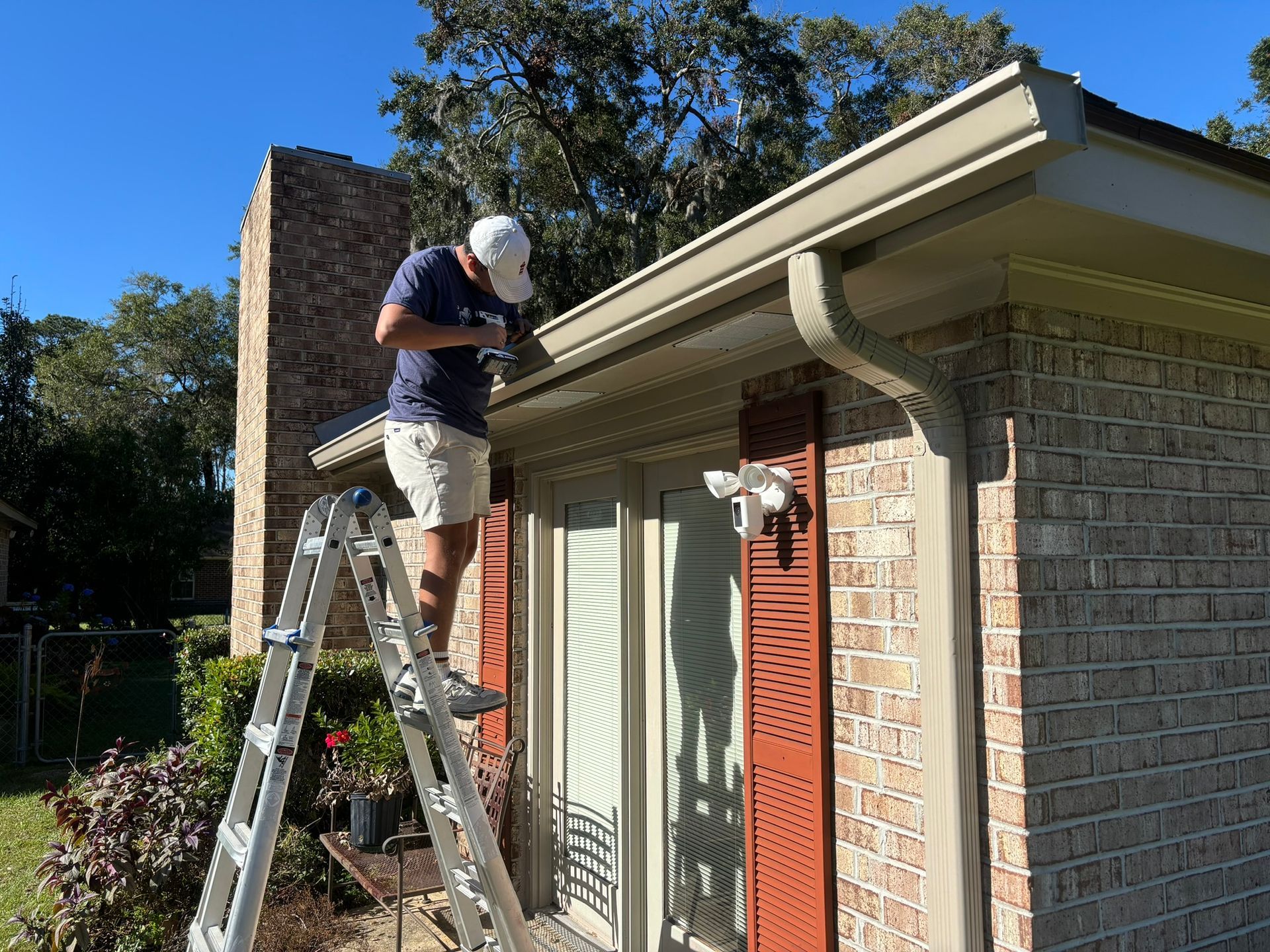 A person standing on a ladder cleans the gutters on the exterior of a brick house on a sunny day.