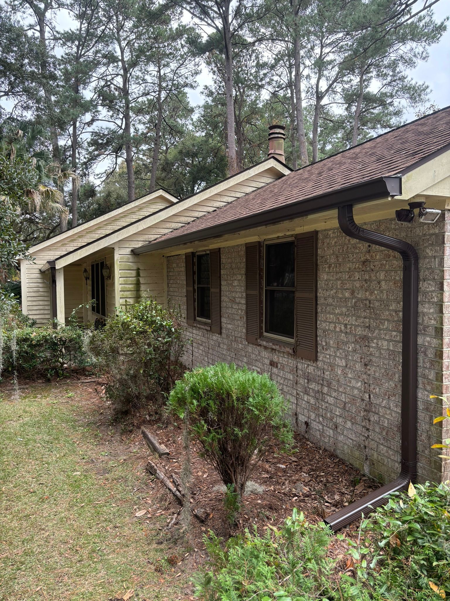 A side view of a brick house with brown shutters, a shingled roof, and a dark brown downspout, set among trees and bushes.