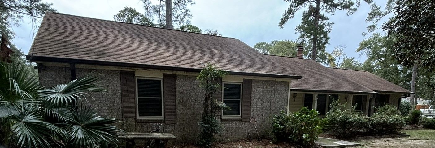 A single-story, stone-exterior house with a brown shingled roof, surrounded by trees and shrubs on a cloudy day.