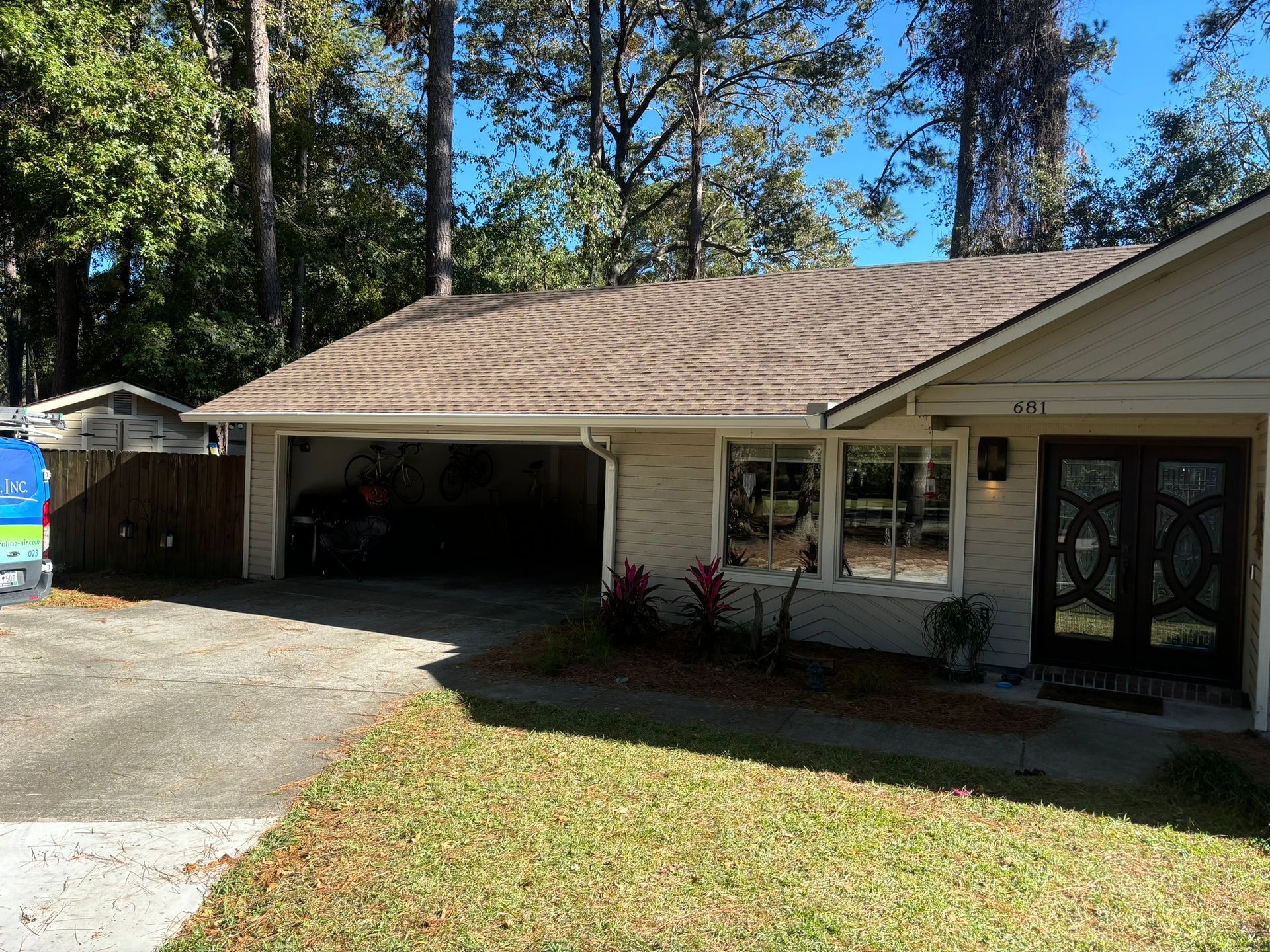A beige single-story house with a two-car open garage, dark front double doors, and a brown shingle roof on a sunny day.