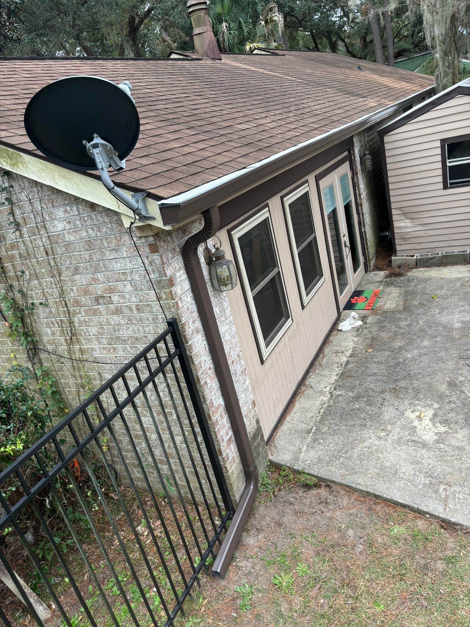 A side view of a house with light-colored stone walls, a brown shingled roof, a satellite dish, and a black metal fence.