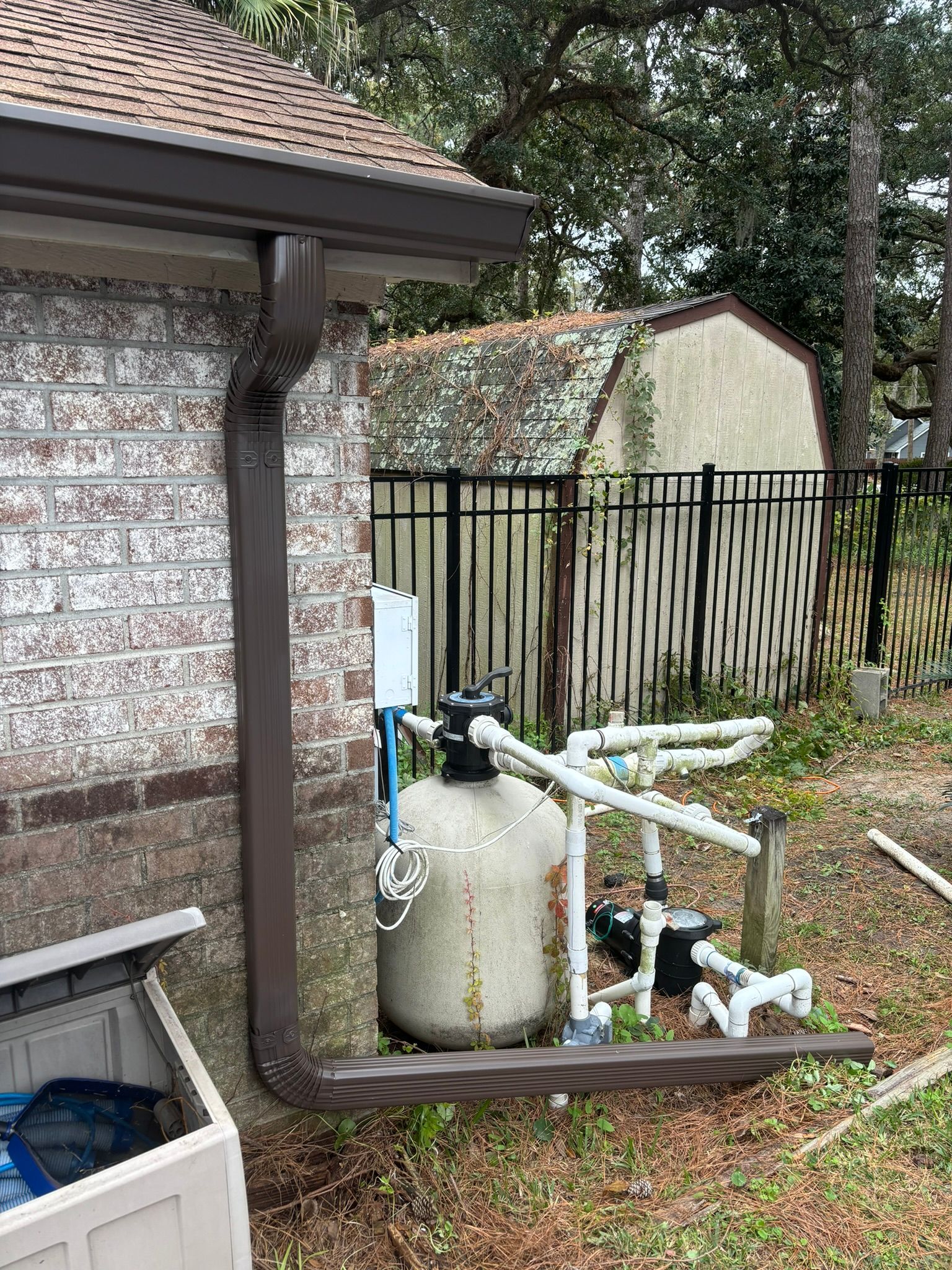 A brown downspout runs along a brick house wall near a pool filter system, piping, and a black metal fence in a yard.