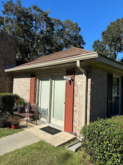 A tan brick, single-story house exterior with brown shutters, a central glass door, and a brown shingled roof under blue sky.