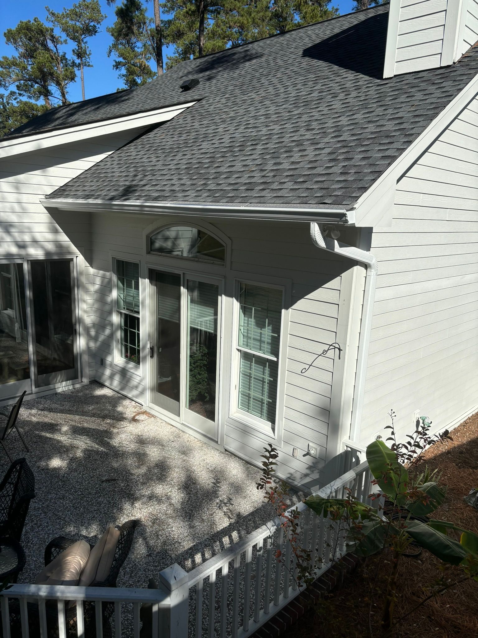A white, one-story house extension with a gray shingled roof, large windows, and a gravel patio area.