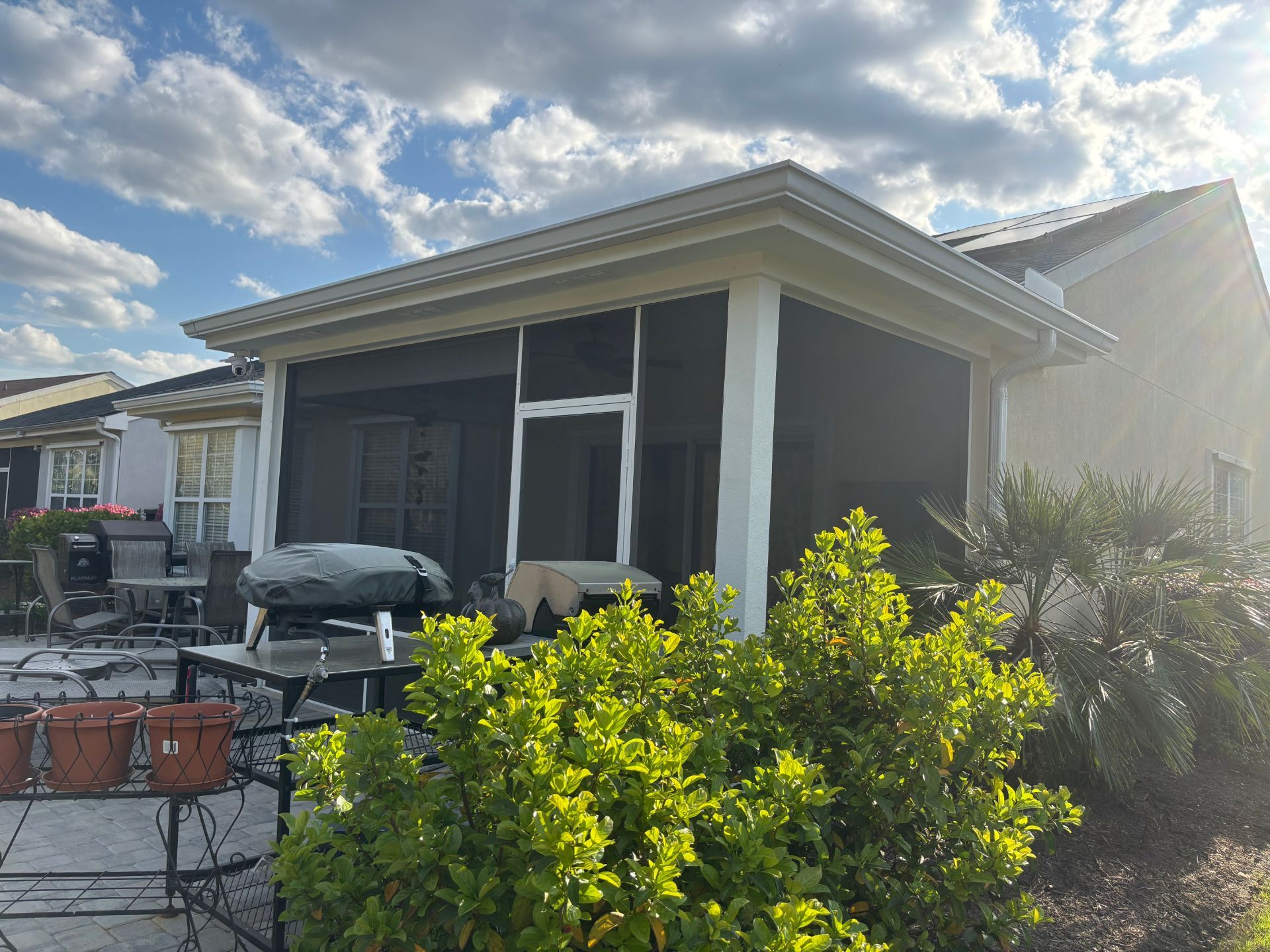 A screened-in back patio with white columns, a grill, and patio furniture, viewed from a sunny backyard with bushes.