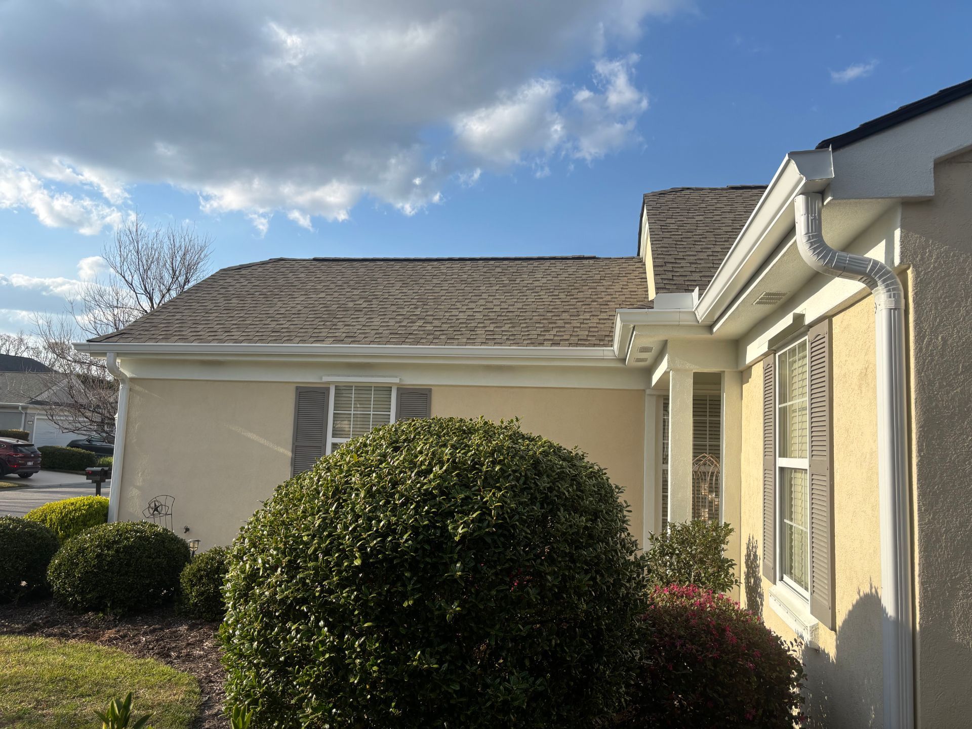 A beige house exterior with a shingled roof, white gutters, and green landscaping under a partly cloudy sky.