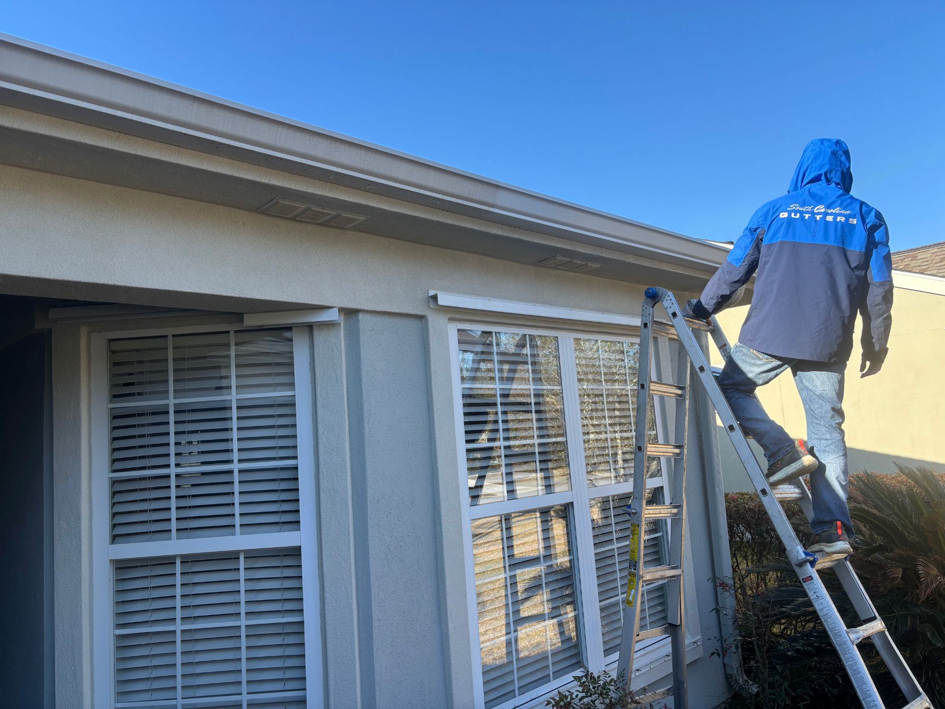 A person in a blue and gray jacket standing on a ladder, working on the exterior of a home near a window.