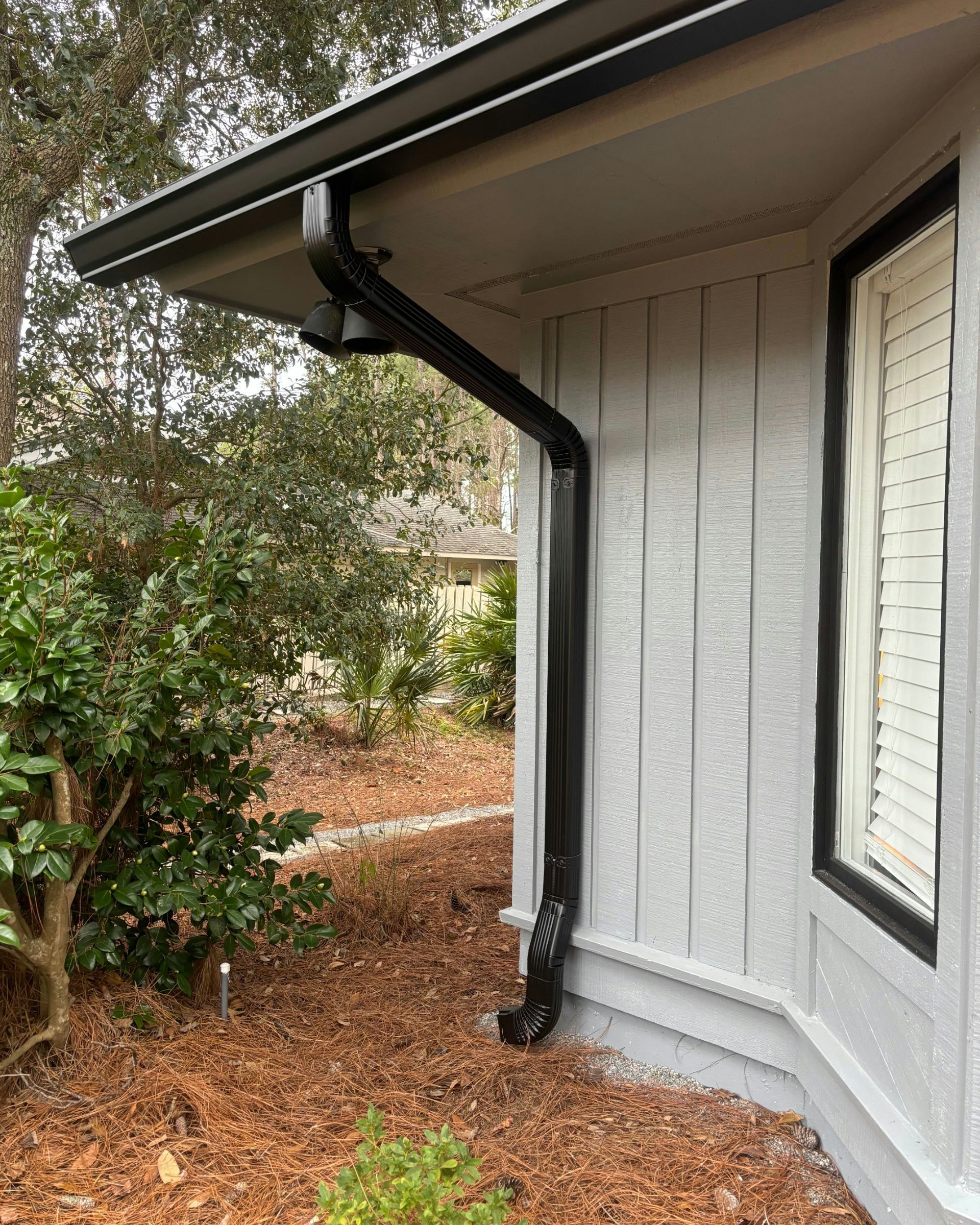 A black gutter downspout attached to the corner of a light grey house with vertical siding and white window shutters.
