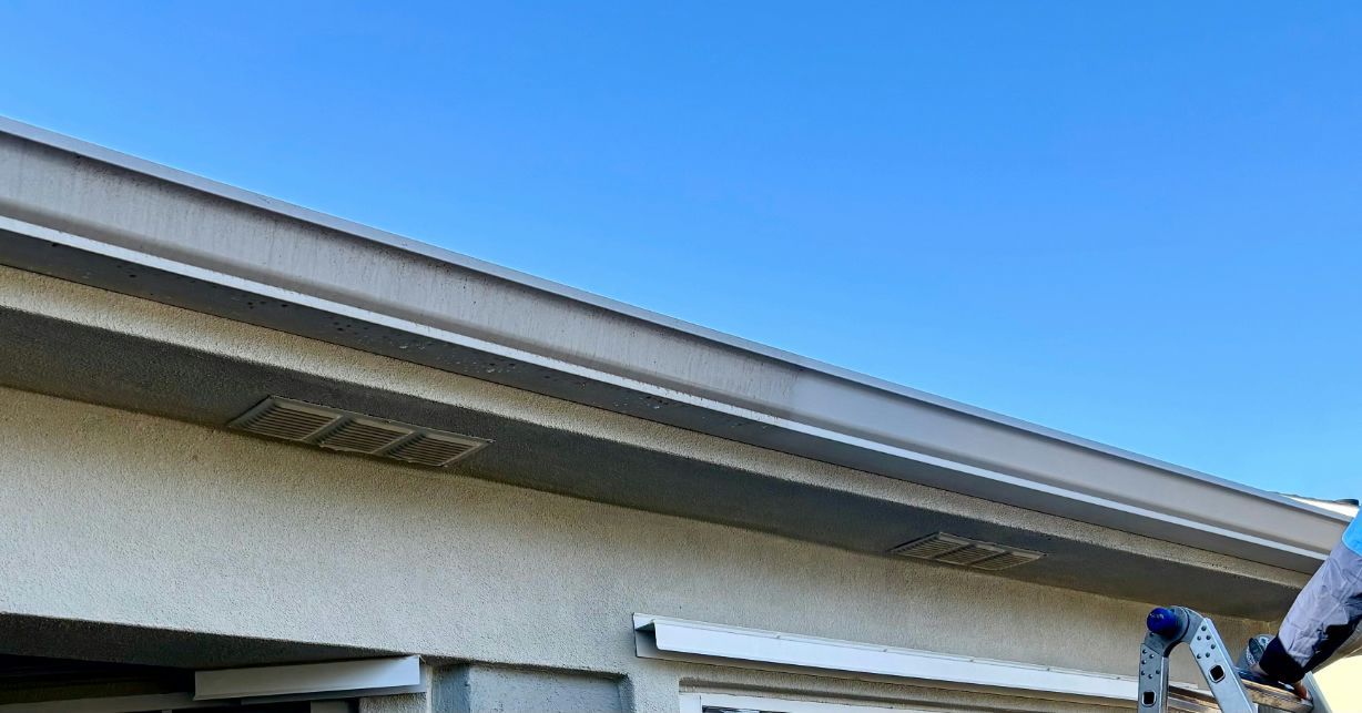 A low-angle view of a grey house exterior with gutters and a soffit vent, partially obscured by a ladder in the foreground.