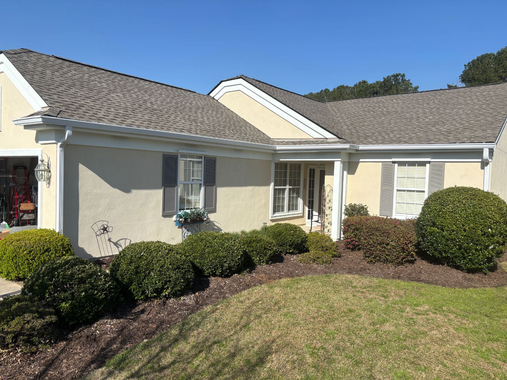 A single-story, beige stucco house with a gray shingled roof, a garage, and landscaped bushes in the front yard.