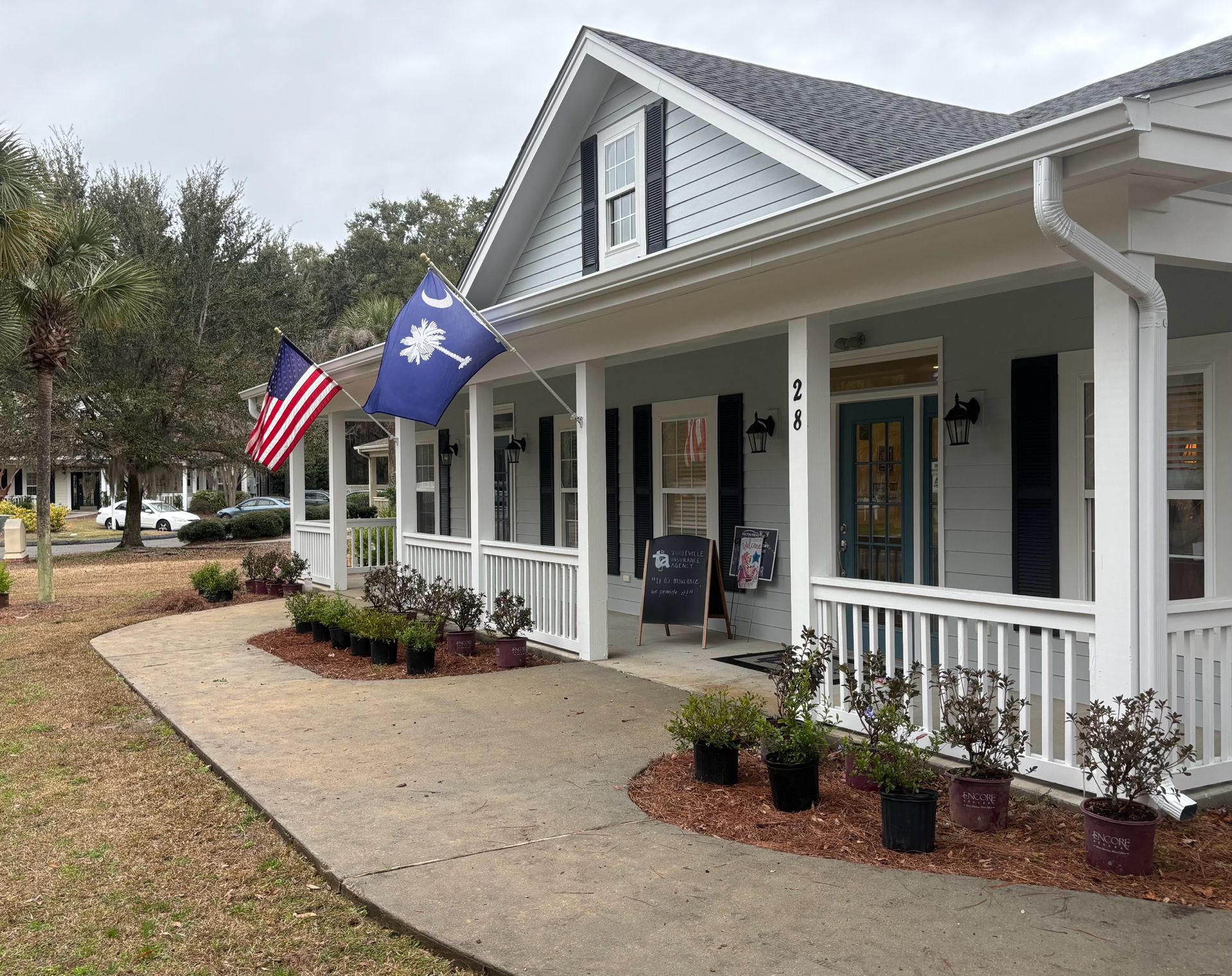 A light blue building with a wrap-around porch, white railings, American and South Carolina flags, and potted shrubs.