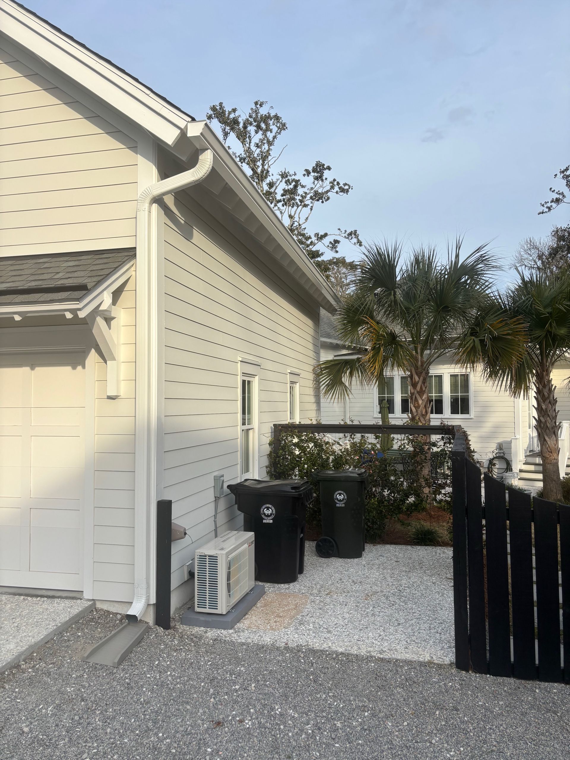 A cream-colored building with a gravel side yard featuring two black trash cans, an AC unit, and a black wooden fence.
