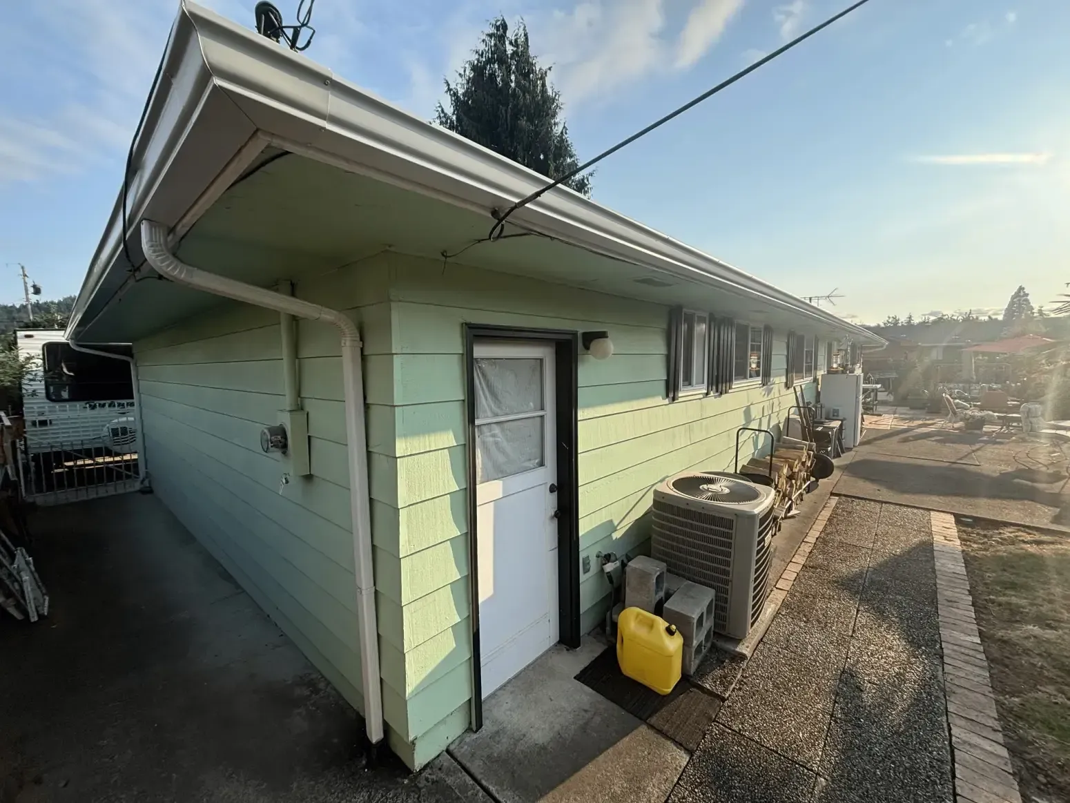 A side view of a pale green house with white trim, a single white door, an AC unit, and a yellow container on the ground.