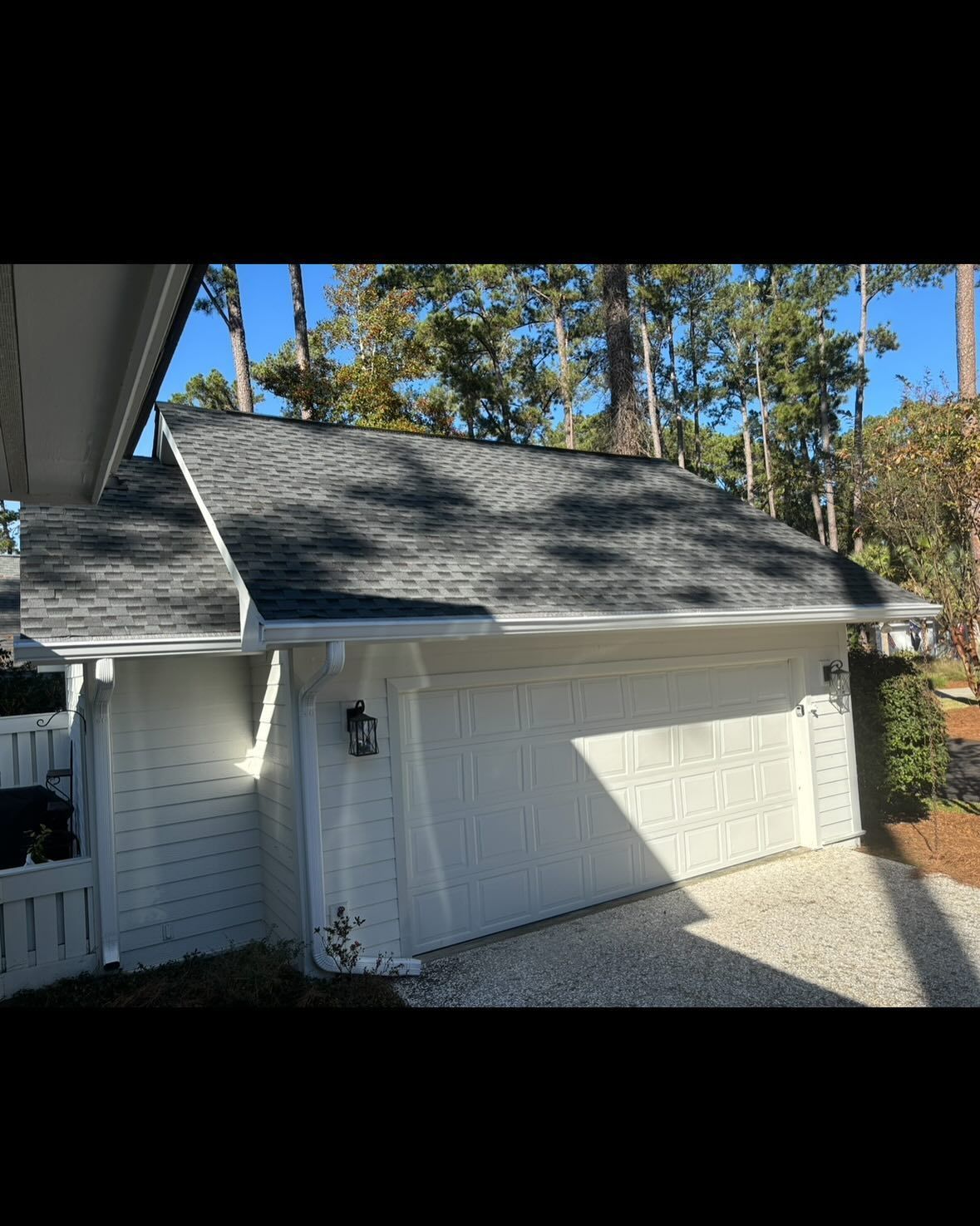 A white, detached garage with a gray shingled roof under a clear blue sky, surrounded by trees.