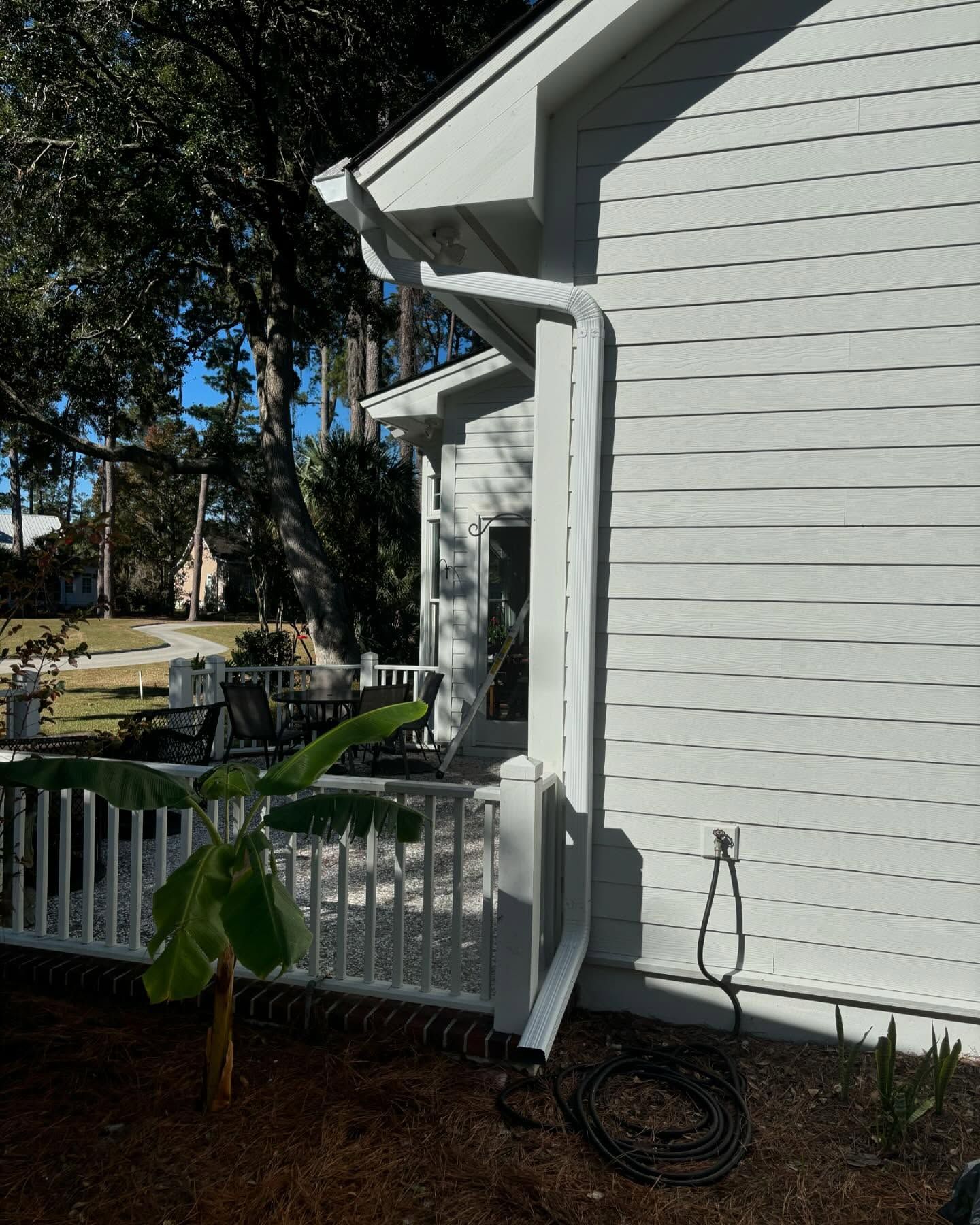 White downspout attached to the side of a white house with a patio and small banana plant in the foreground.