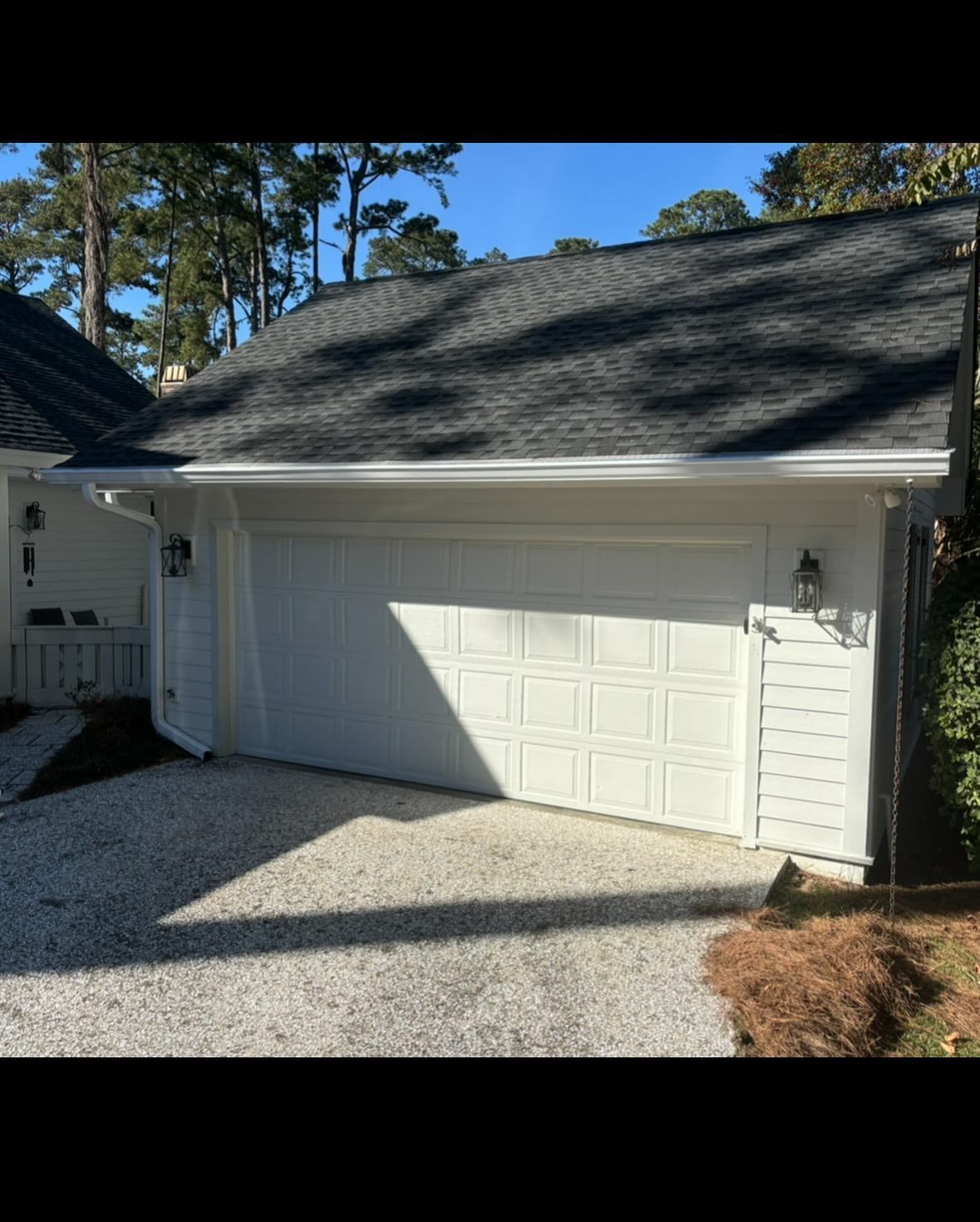 A white, single-car garage with a gray shingled roof, set against a backdrop of trees and a gravel driveway.