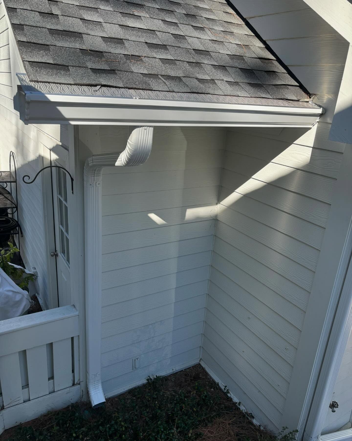 A white exterior wall of a house with a white downspout attached to a white rain gutter under an asphalt shingle roof.