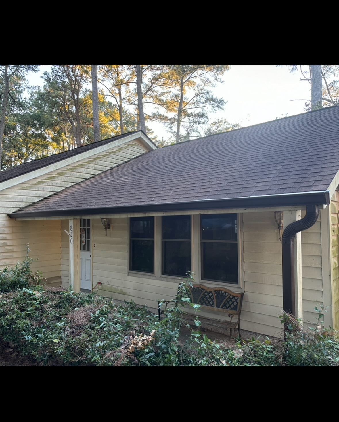 A beige, single-story house exterior with a dark shingled roof, three windows, a door, and a garden in the foreground.