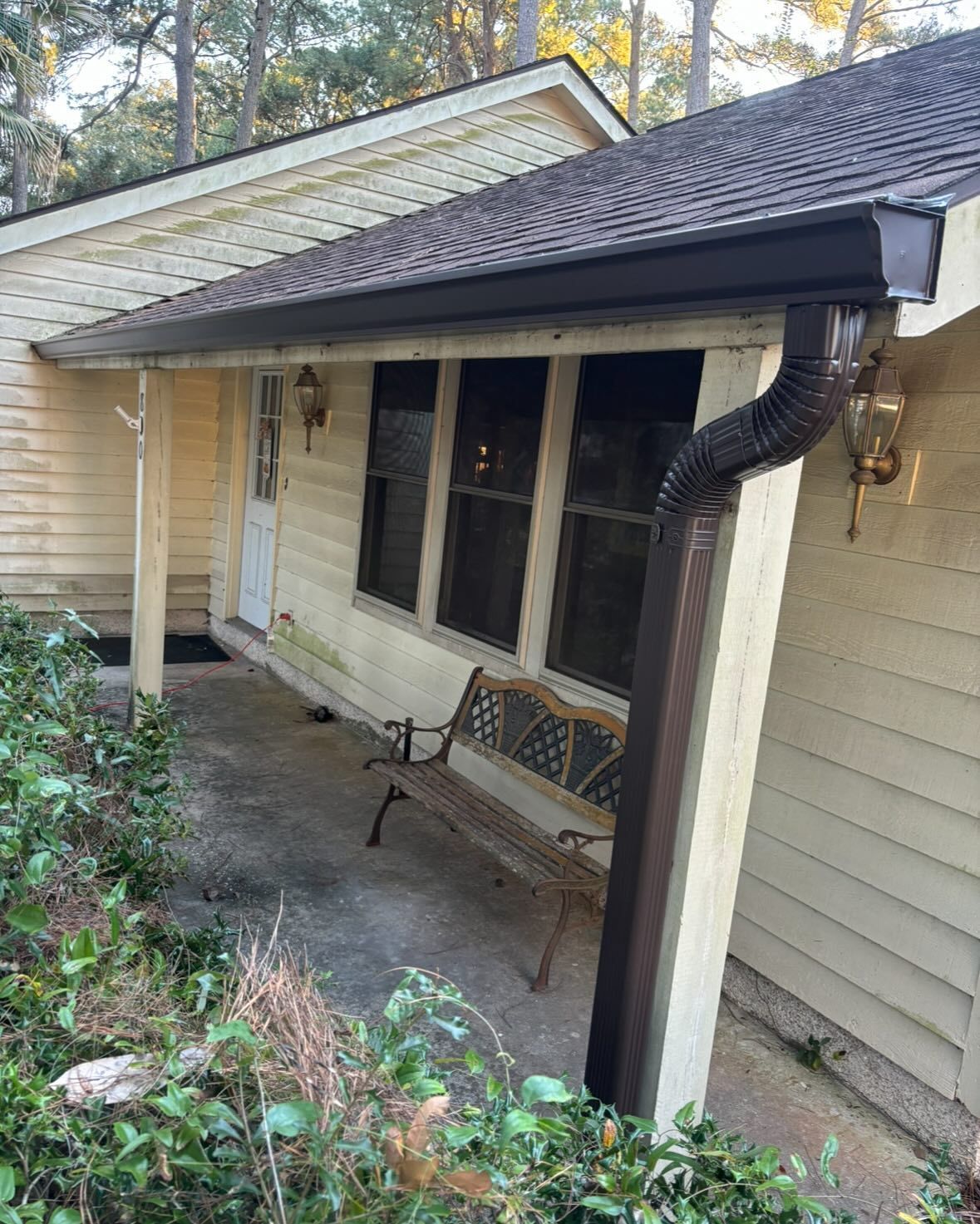 A beige house with a covered porch featuring a brown metal bench, dark window frames, and a brown gutter downspout.