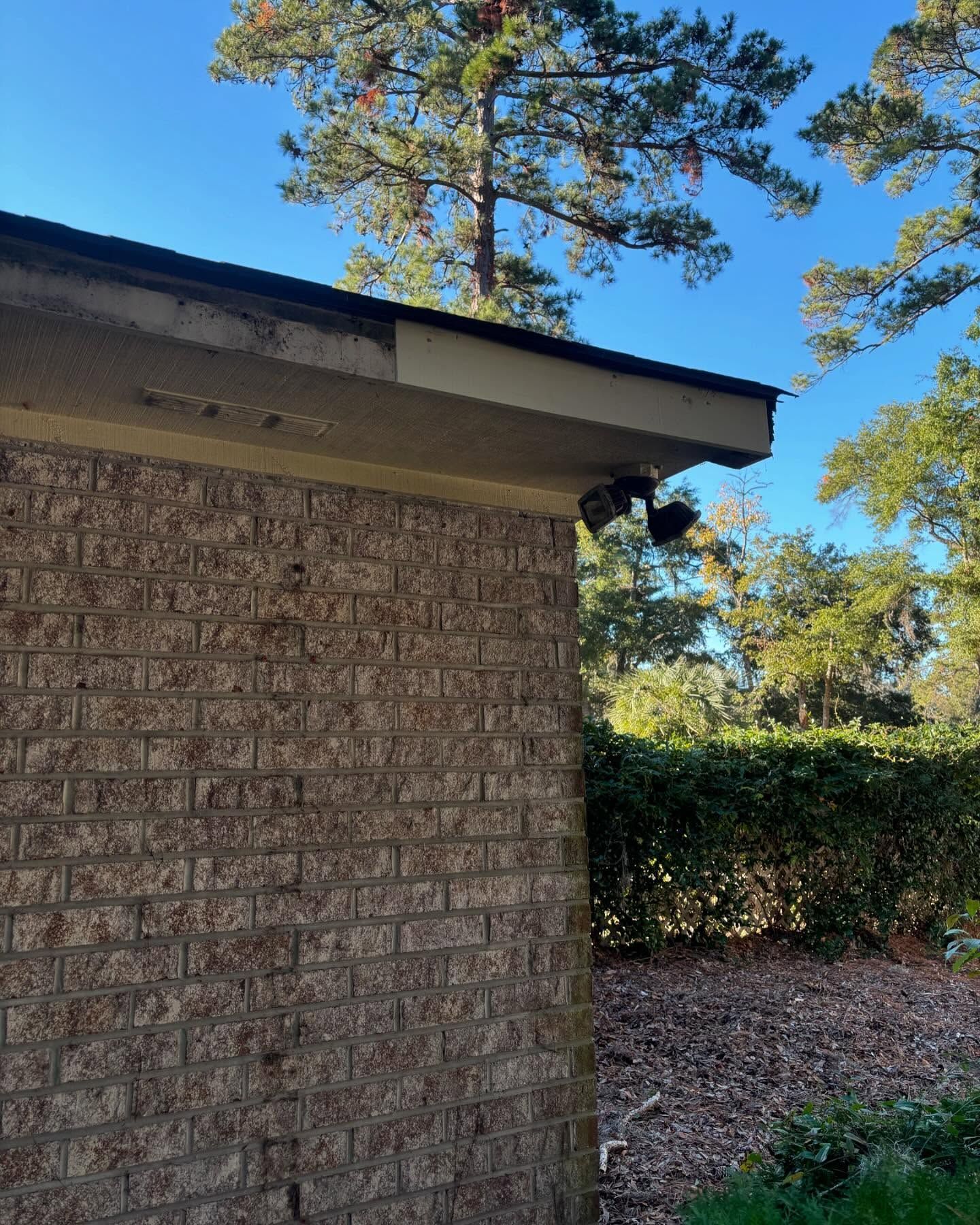 An outdoor view of a brick house corner with a tan soffit, a dark dual floodlight fixture, and tall trees in the background.