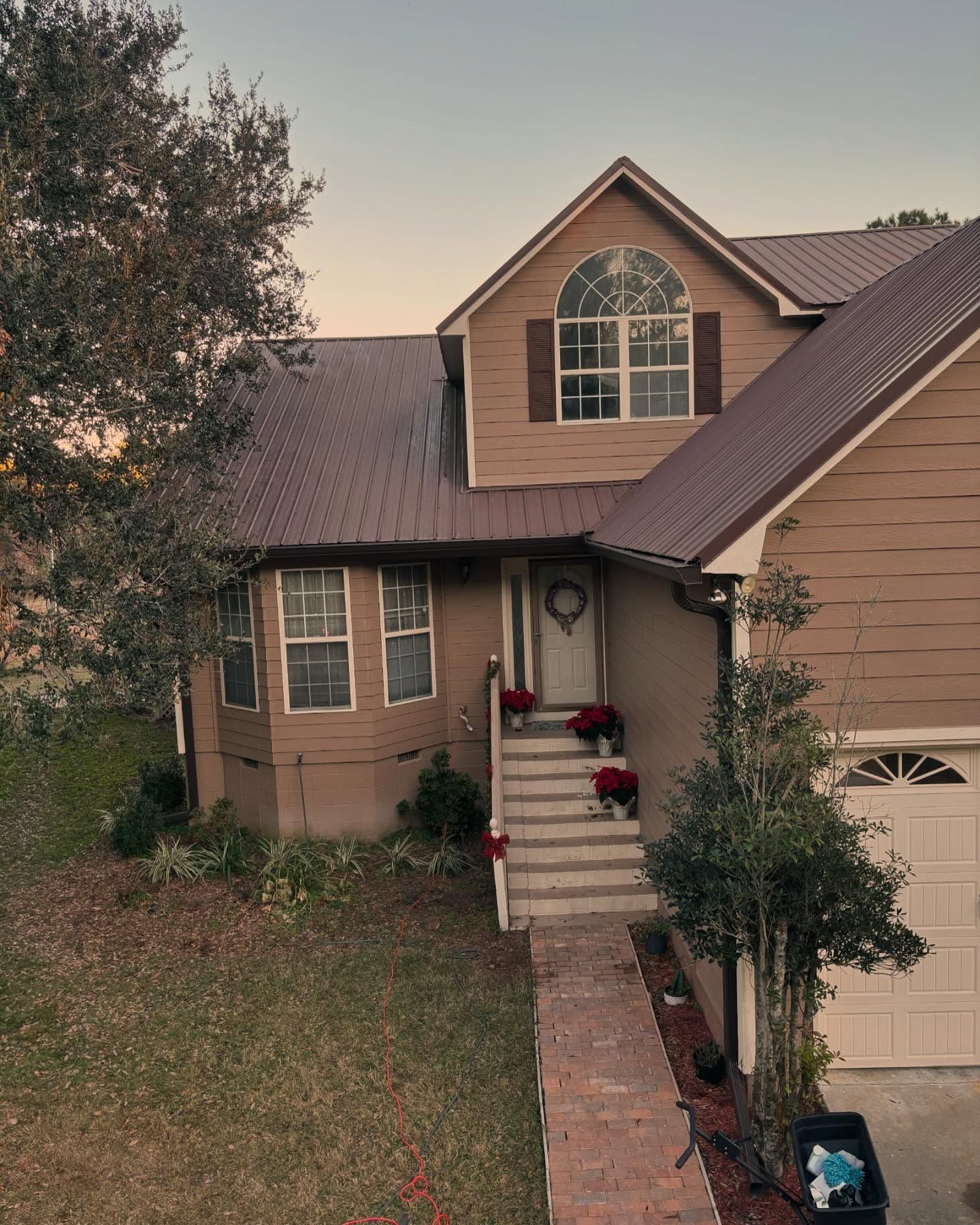 A two-story tan house with a dark metal roof, brick walkway, and a garage under a soft, golden-hour sky.