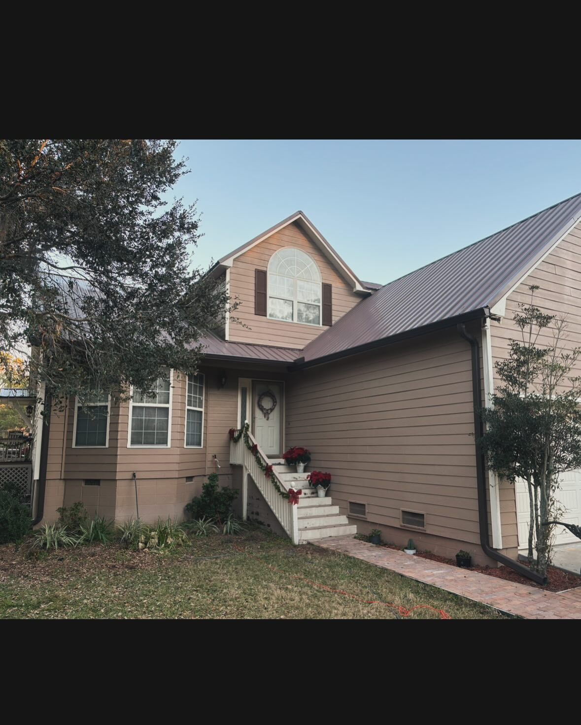 A two-story tan house with a dark metal roof, a front porch with stairs, and a sidewalk leading to the entrance.