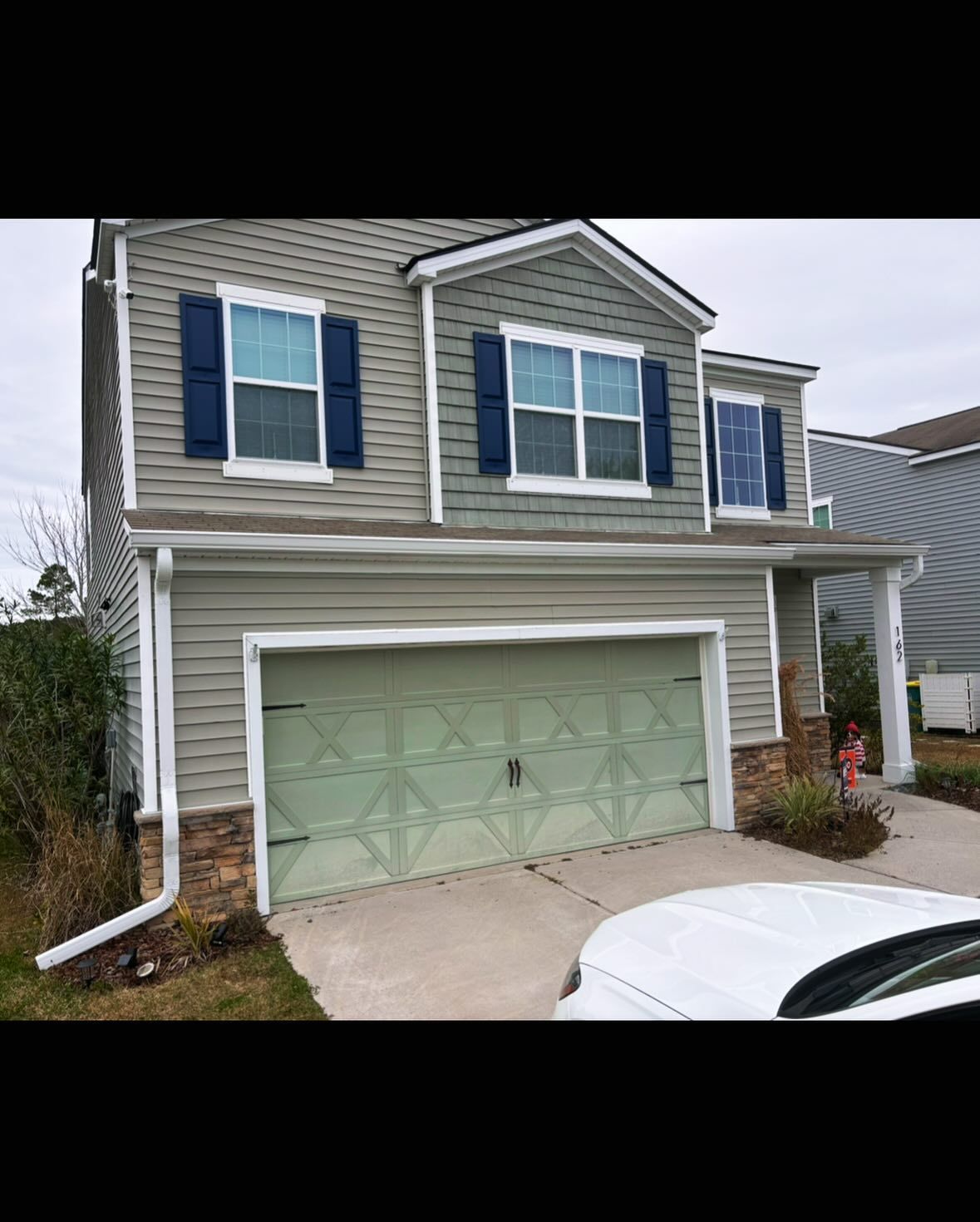 Two-story suburban house with beige siding, dark blue shutters, stone accents, and a light green garage door.