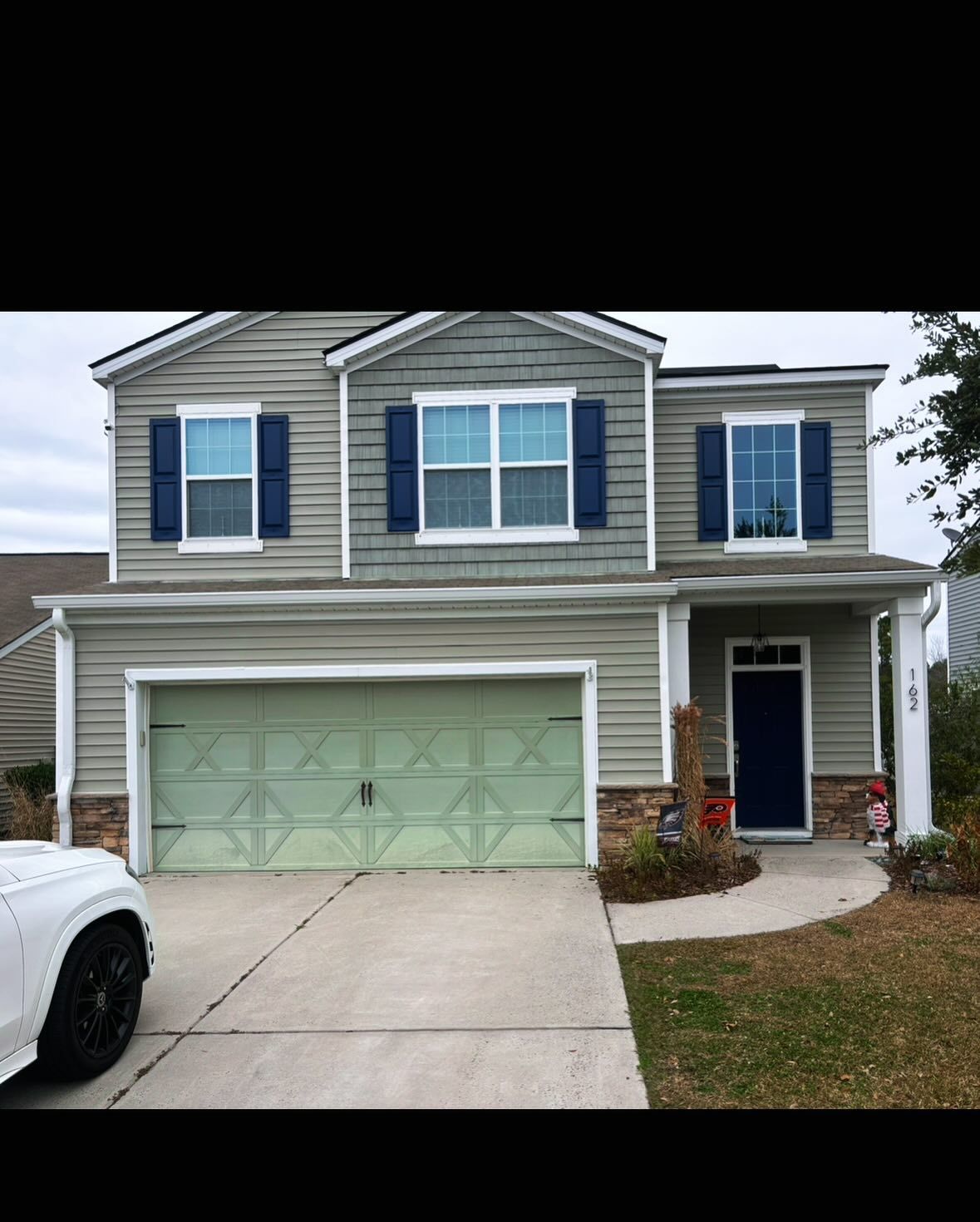 A two-story suburban house with light gray siding, a light green garage door, blue shutters, and a dark blue front door.