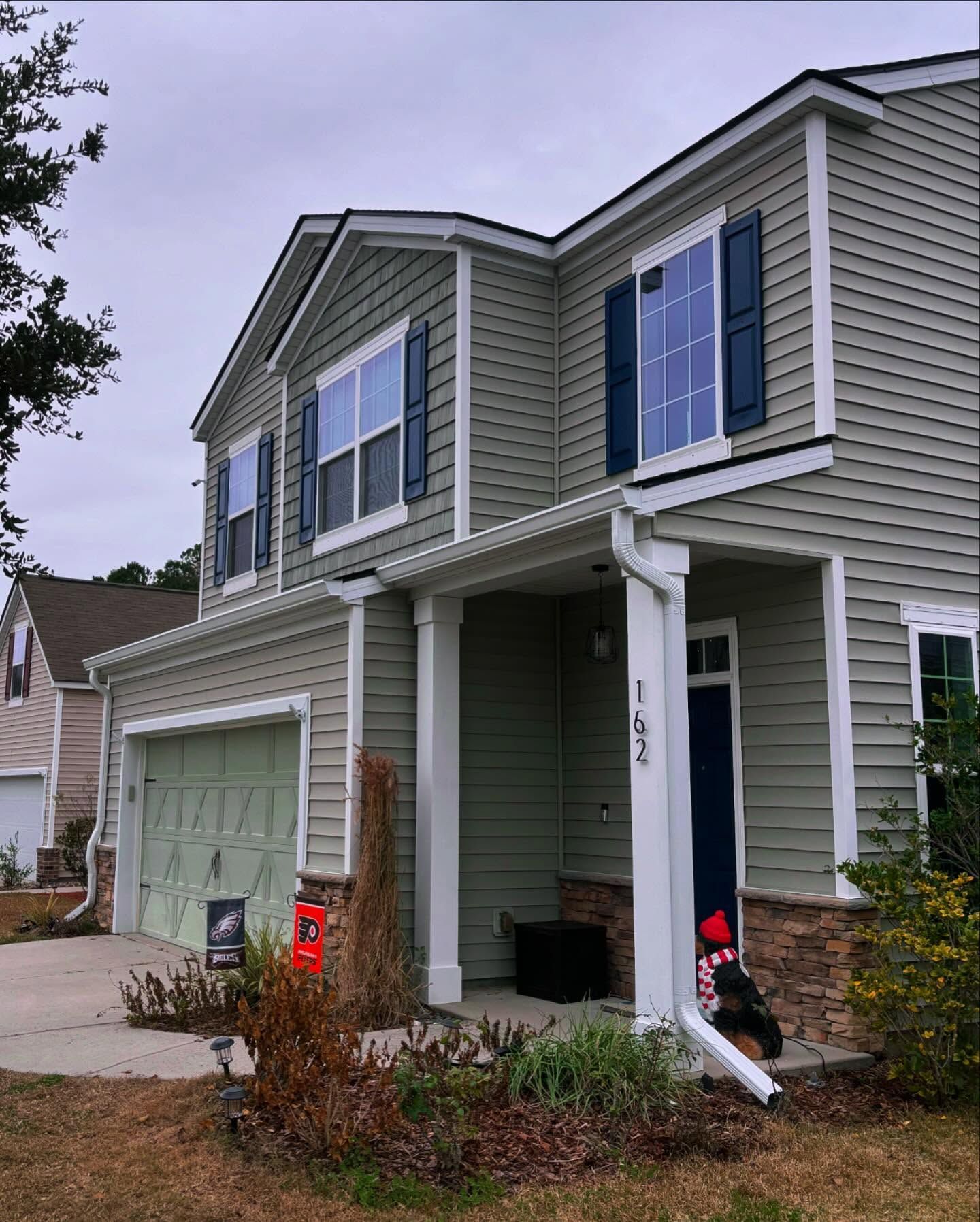 A two-story, green-sided house with a garage, front porch, and blue window shutters on an overcast day.