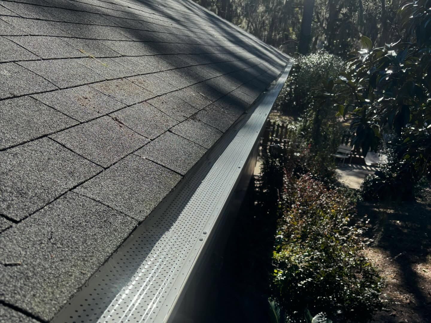 A shingled roof edge with a metal gutter guard installed, seen from a high angle next to trees.