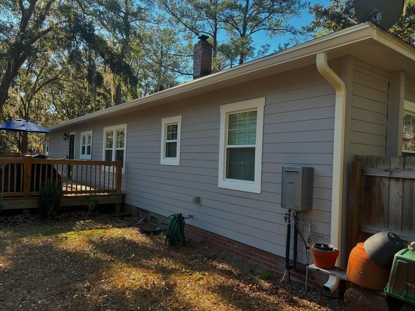 A side view of a gray-sided house with white window frames, a wooden deck, a brick foundation, and a chimney.