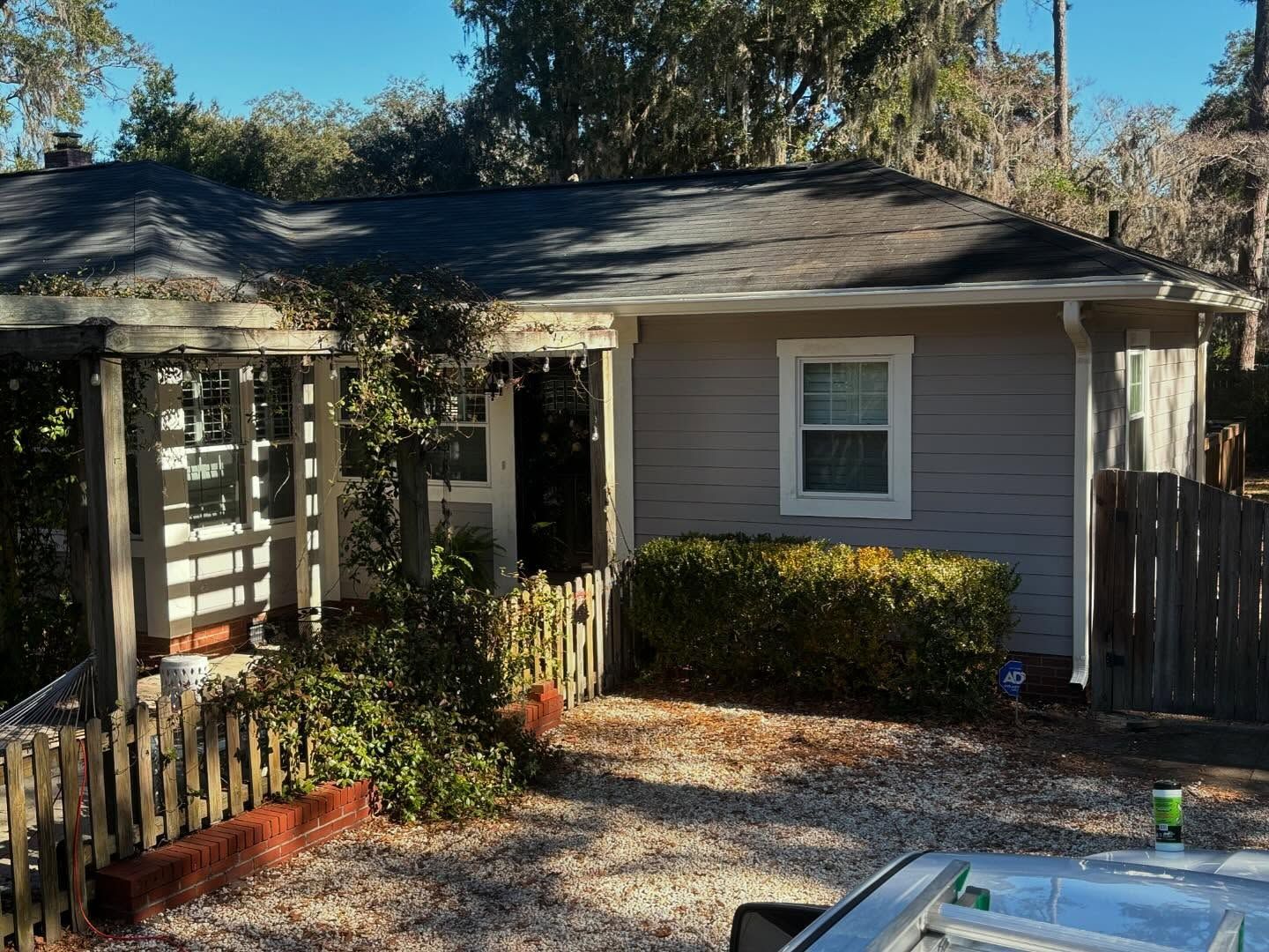 A one-story gray house with a roofed porch, surrounded by trees and enclosed by a wooden fence in a gravel yard.